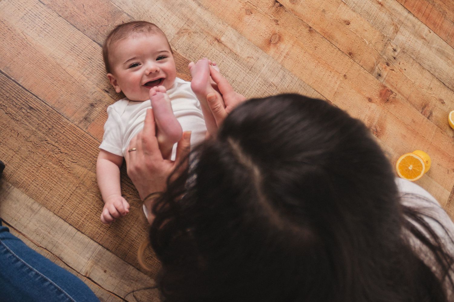 A smiling baby lying on a wooden floor while a mother plays with them during a photoshoot.