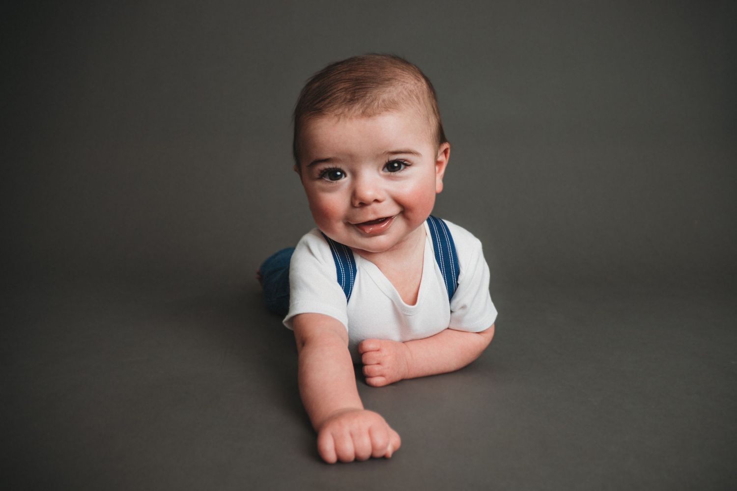 A happy baby boy wearing white onesie and blue suspenders smiling while lying on a gray studio floor.