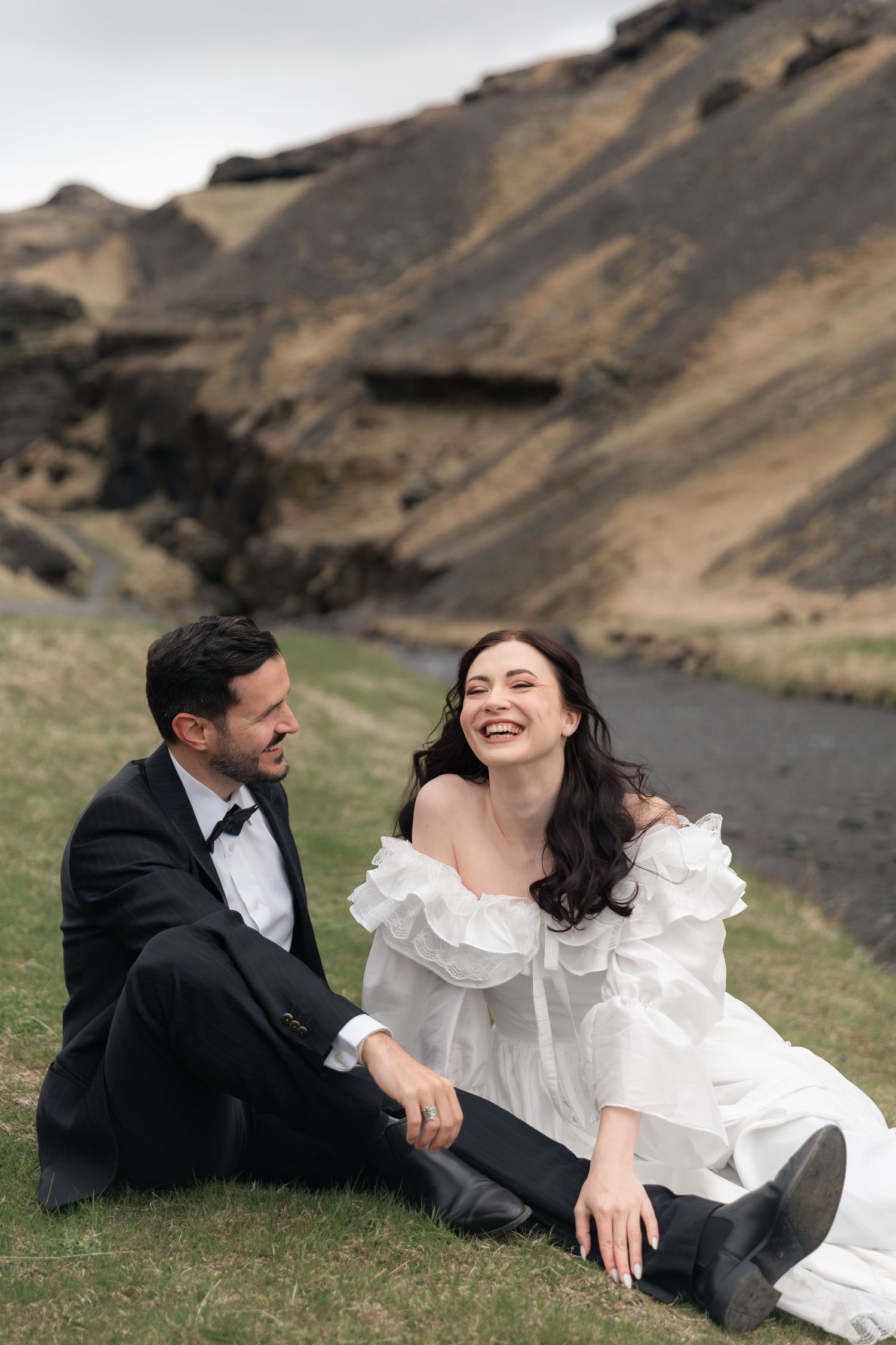 A joyful bride in a white ruffled gown and groom in black tuxedo sit laughing on grass with dramatic mountains behind.