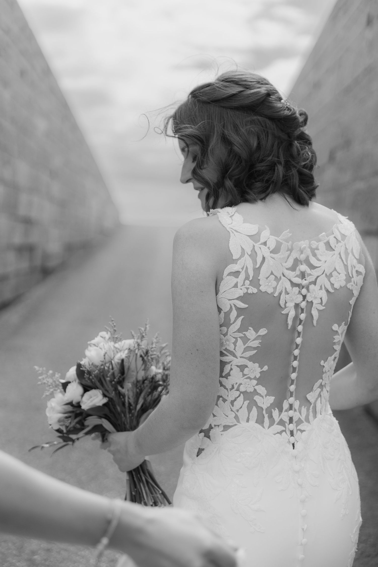 Black and white photo of bride holding bouquet, showing intricate lace back detail of wedding dress.