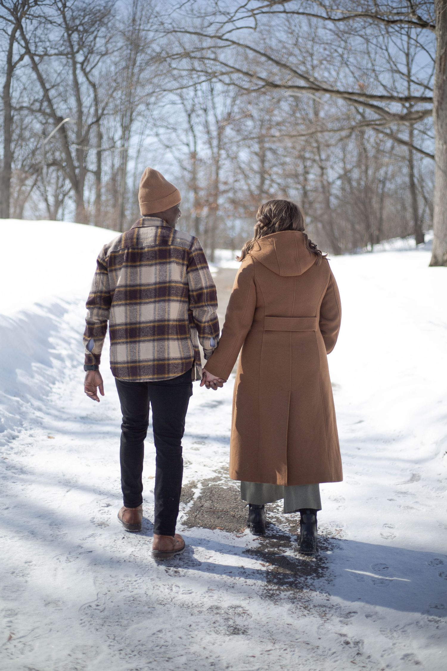 A couple walks hand in hand down a snowy path through bare winter trees.