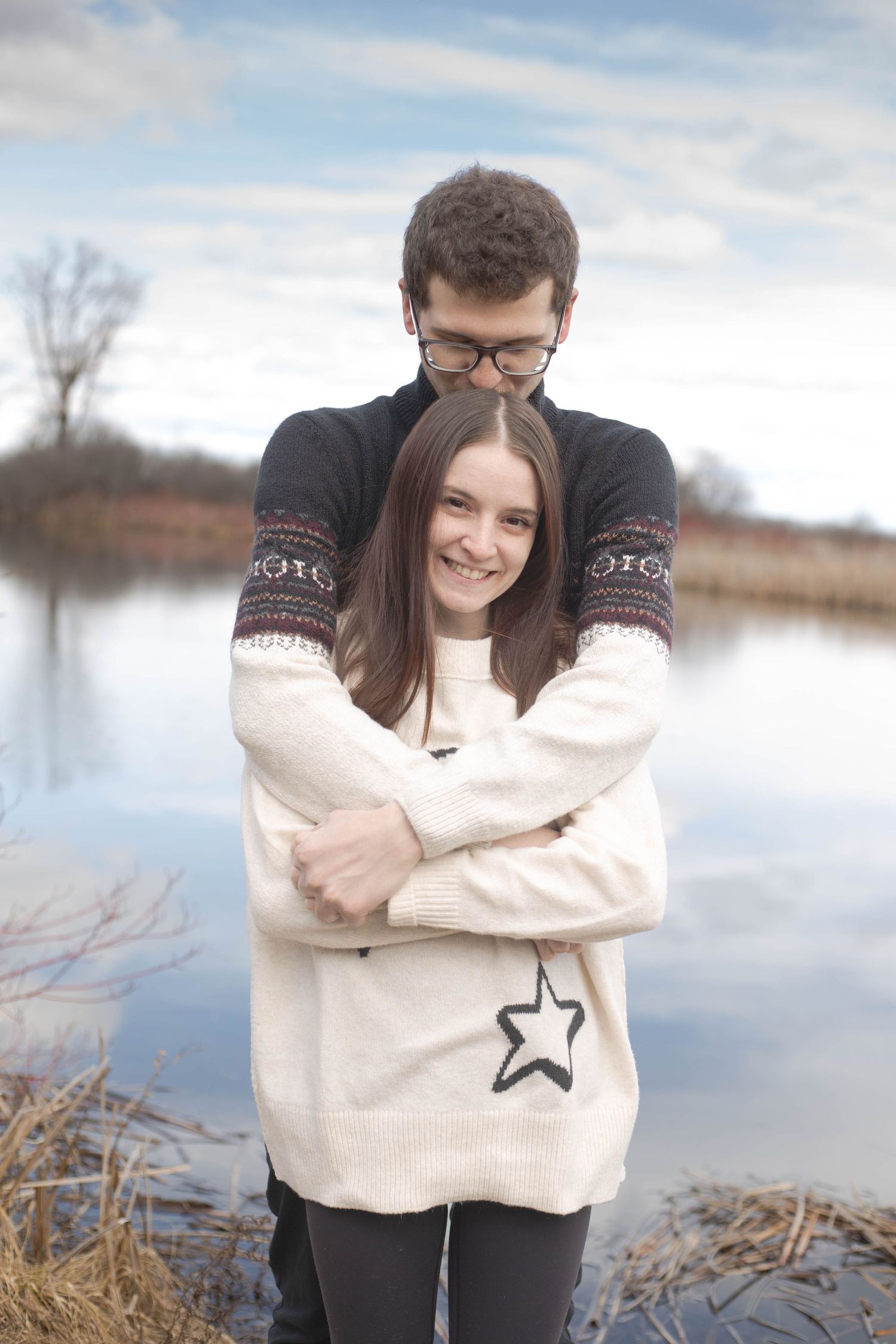 A couple embraces by a serene lake on an overcast day, wearing cozy winter sweaters in a romantic outdoor setting.