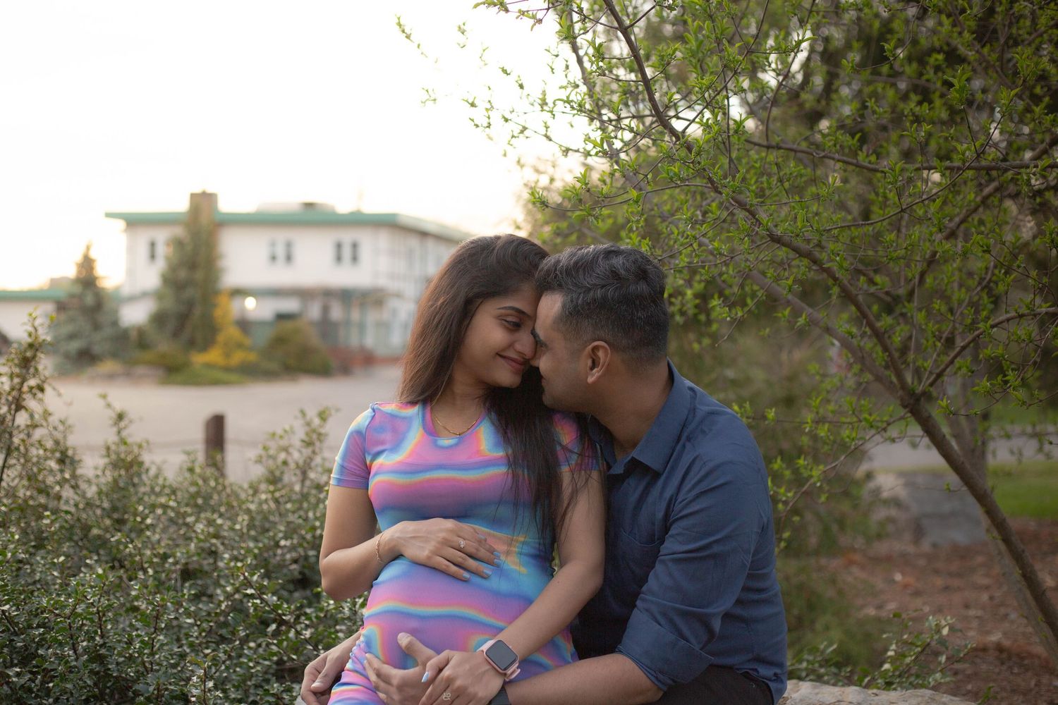 A couple sits together outdoors during a romantic sunset moment in a garden setting with houses in the background.