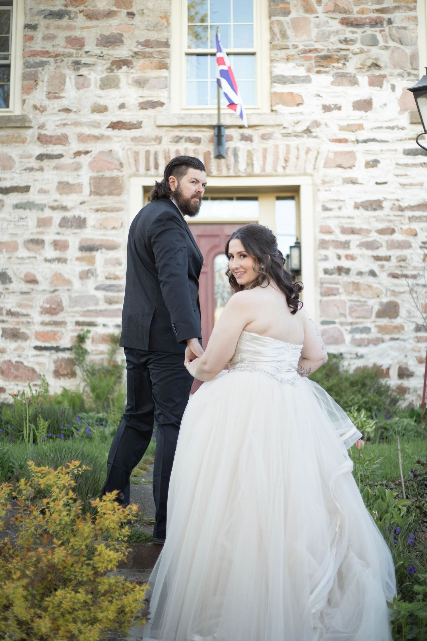 A wedding couple stands outside a rustic stone church, with the bride wearing a flowing white ballgown dress.