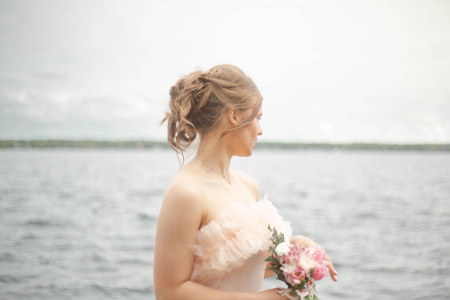 A bride in a strapless gown holds a pink bouquet while looking out over a serene lake on her wedding day.