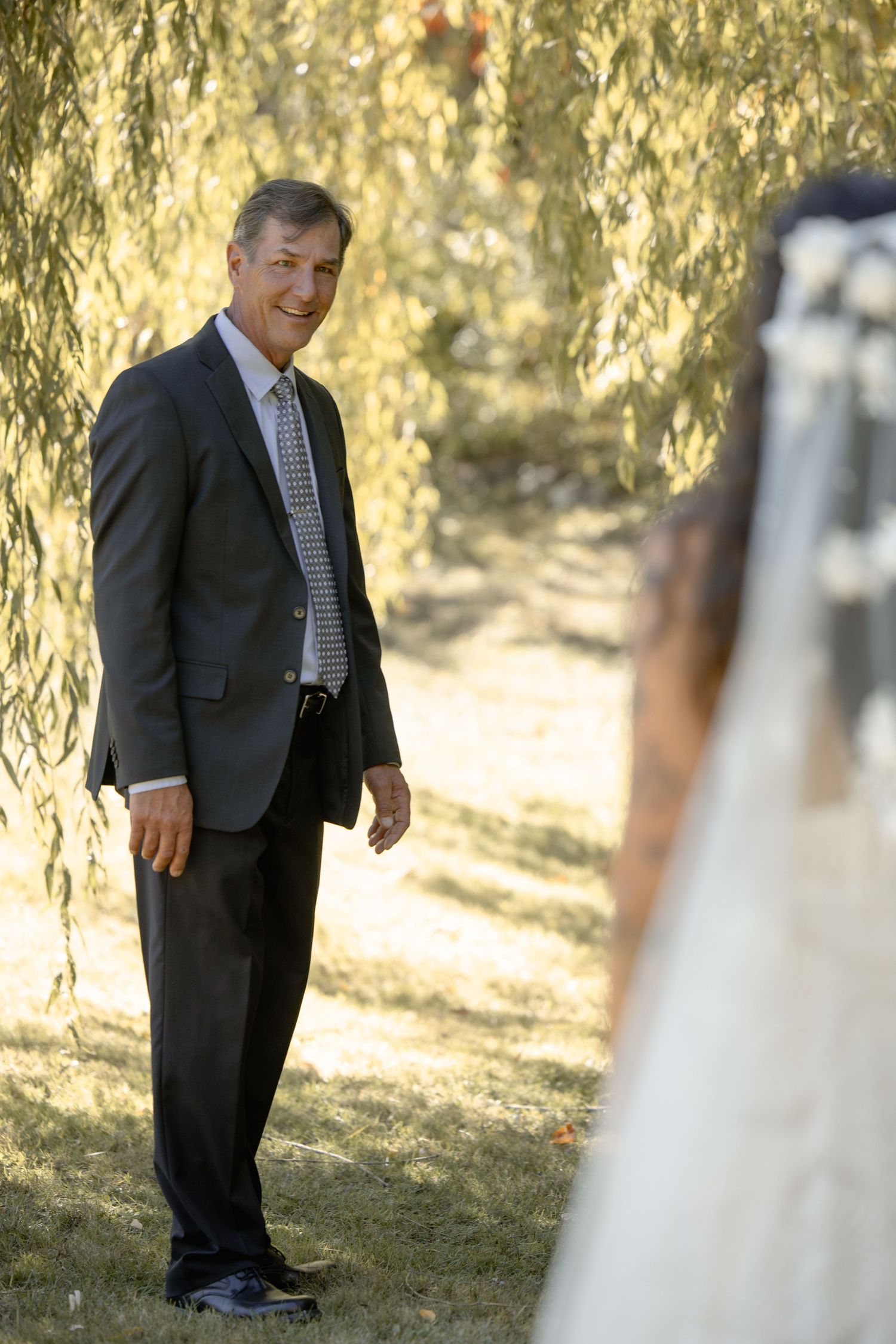 A person in a dark suit and tie stands under a willow tree during what appears to be a wedding photo session.