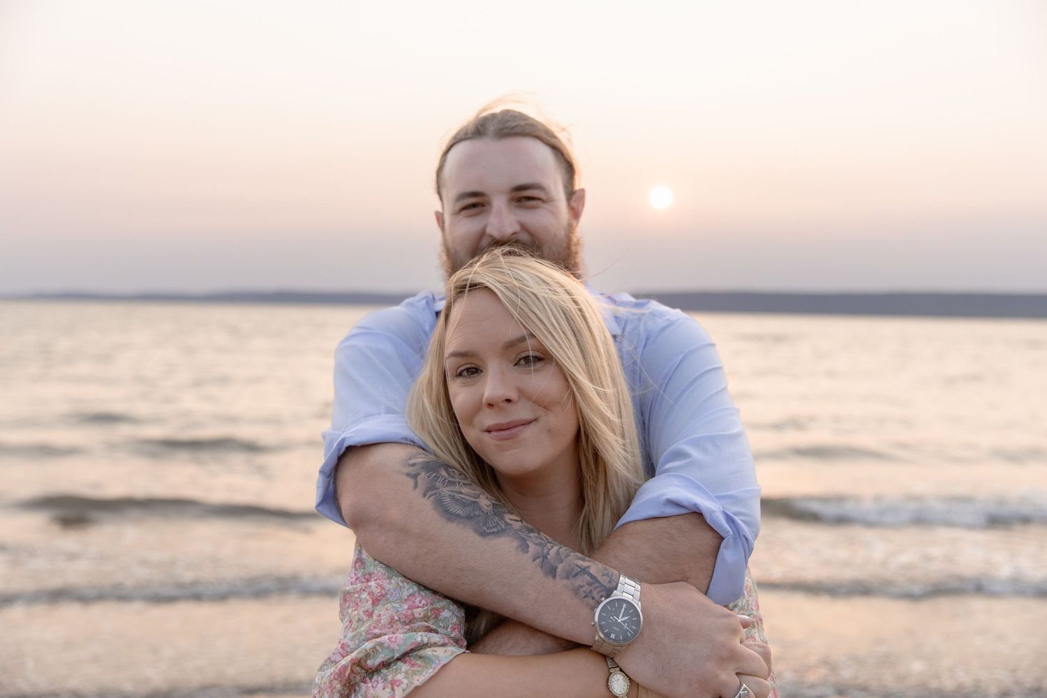 Couple embraces while sitting on beach during sunset session with ocean waves in background.