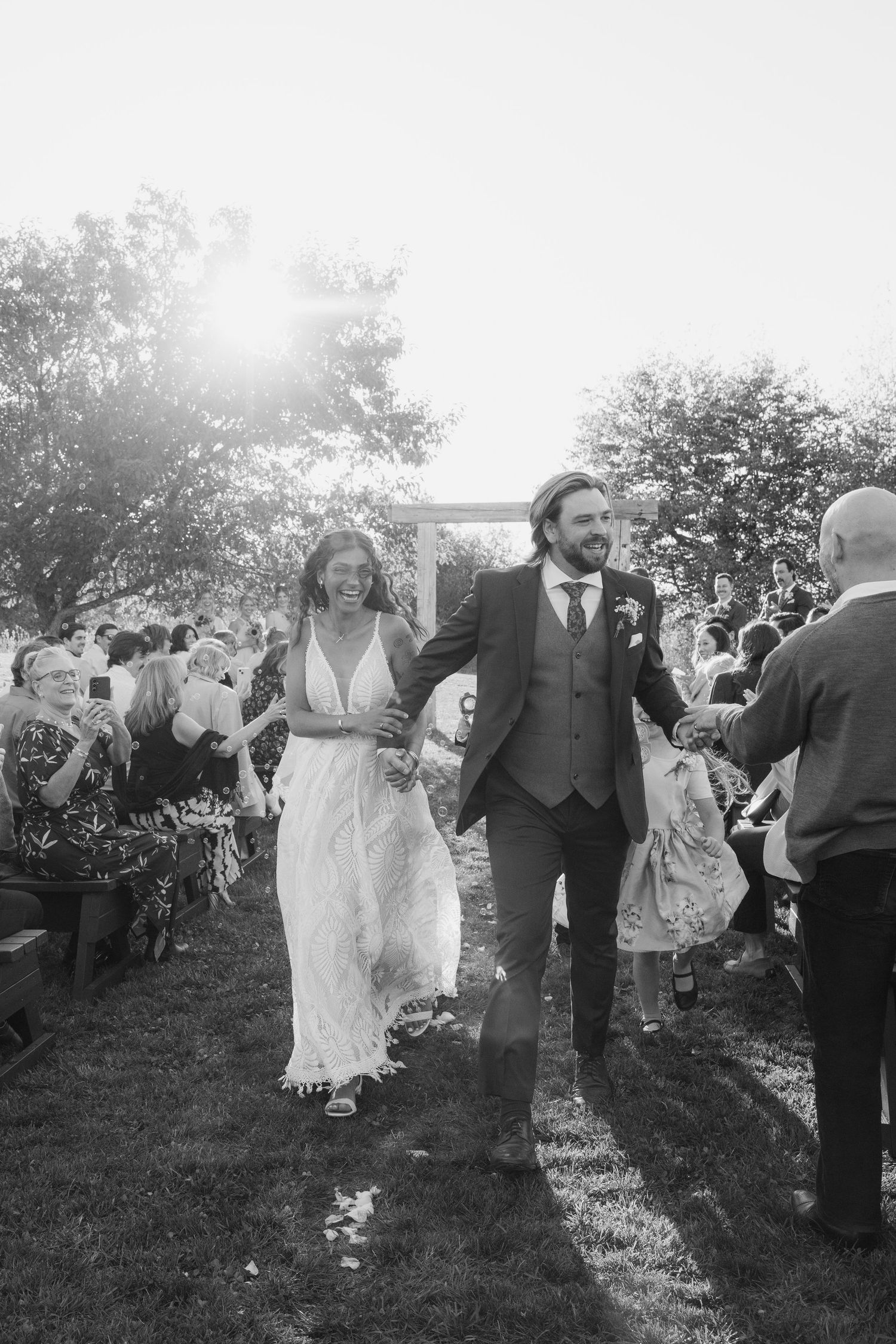 A joyful couple walks hand in hand through their outdoor wedding reception as sunlight streams through the trees.