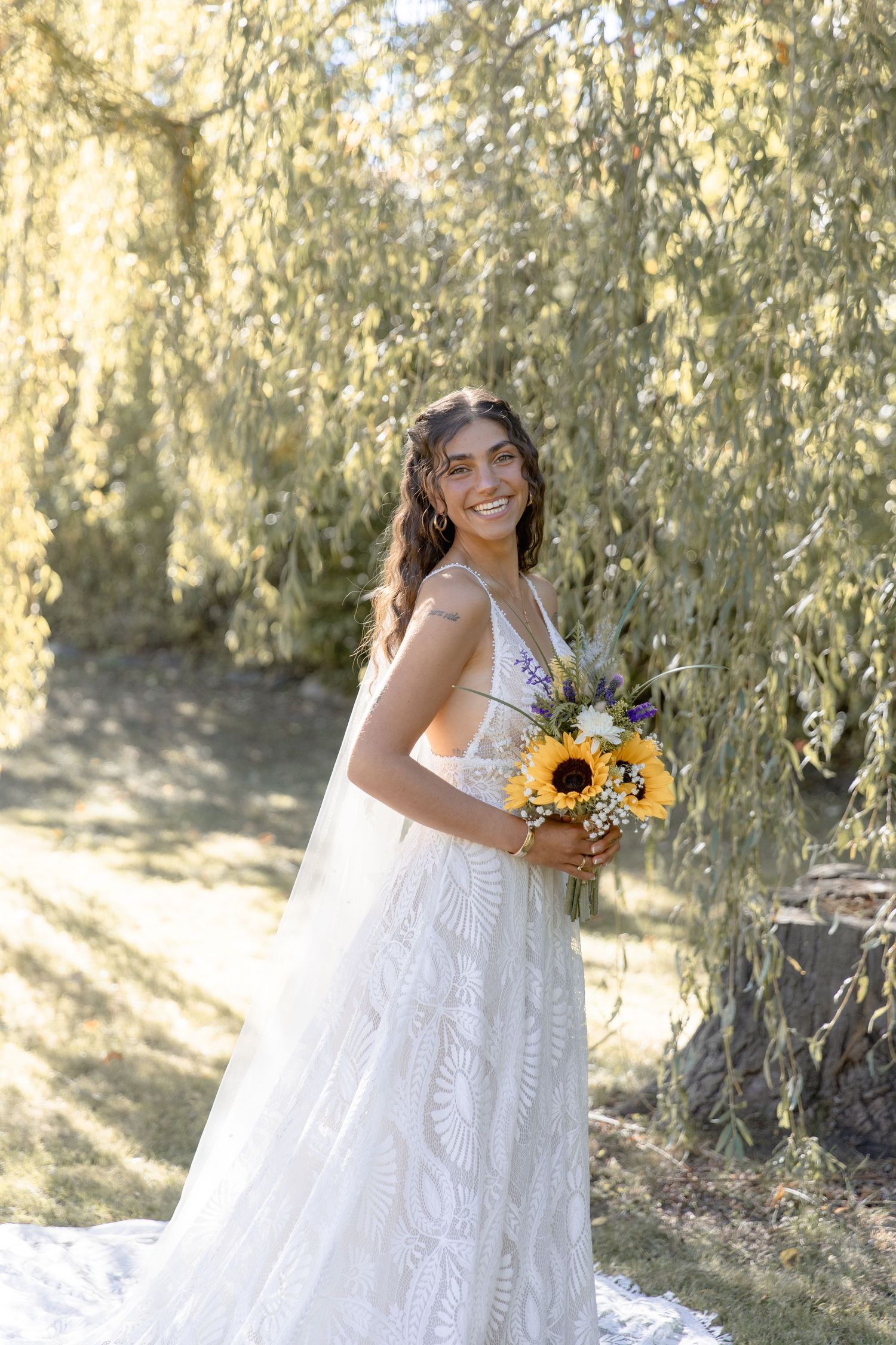 A young girl in a white lace dress poses outdoors with sunflowers against a natural willow tree backdrop.