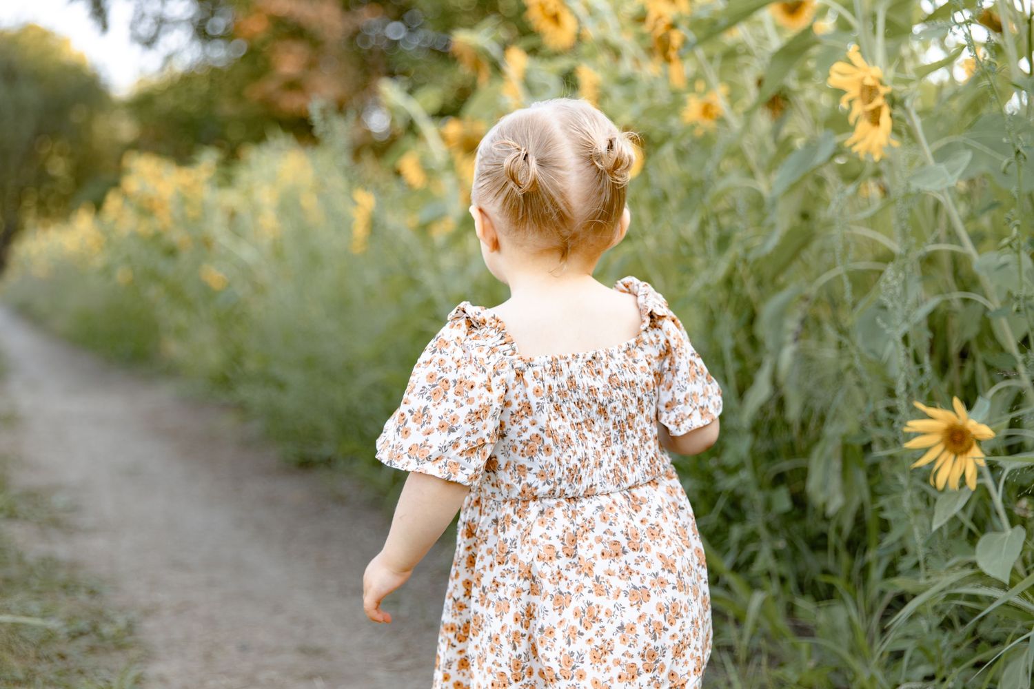 A toddler in a floral dress explores a garden path lined with tall yellow flowers and greenery.
