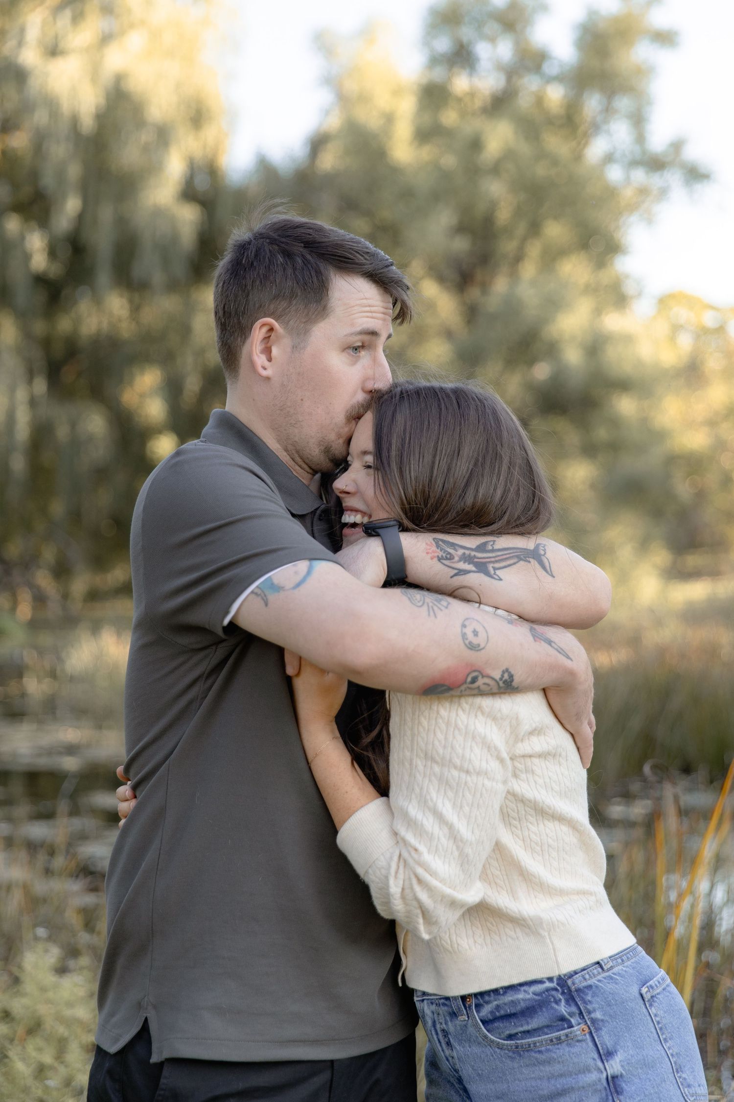 A couple embraces tenderly in an outdoor setting with golden sunlight filtering through trees in the background.