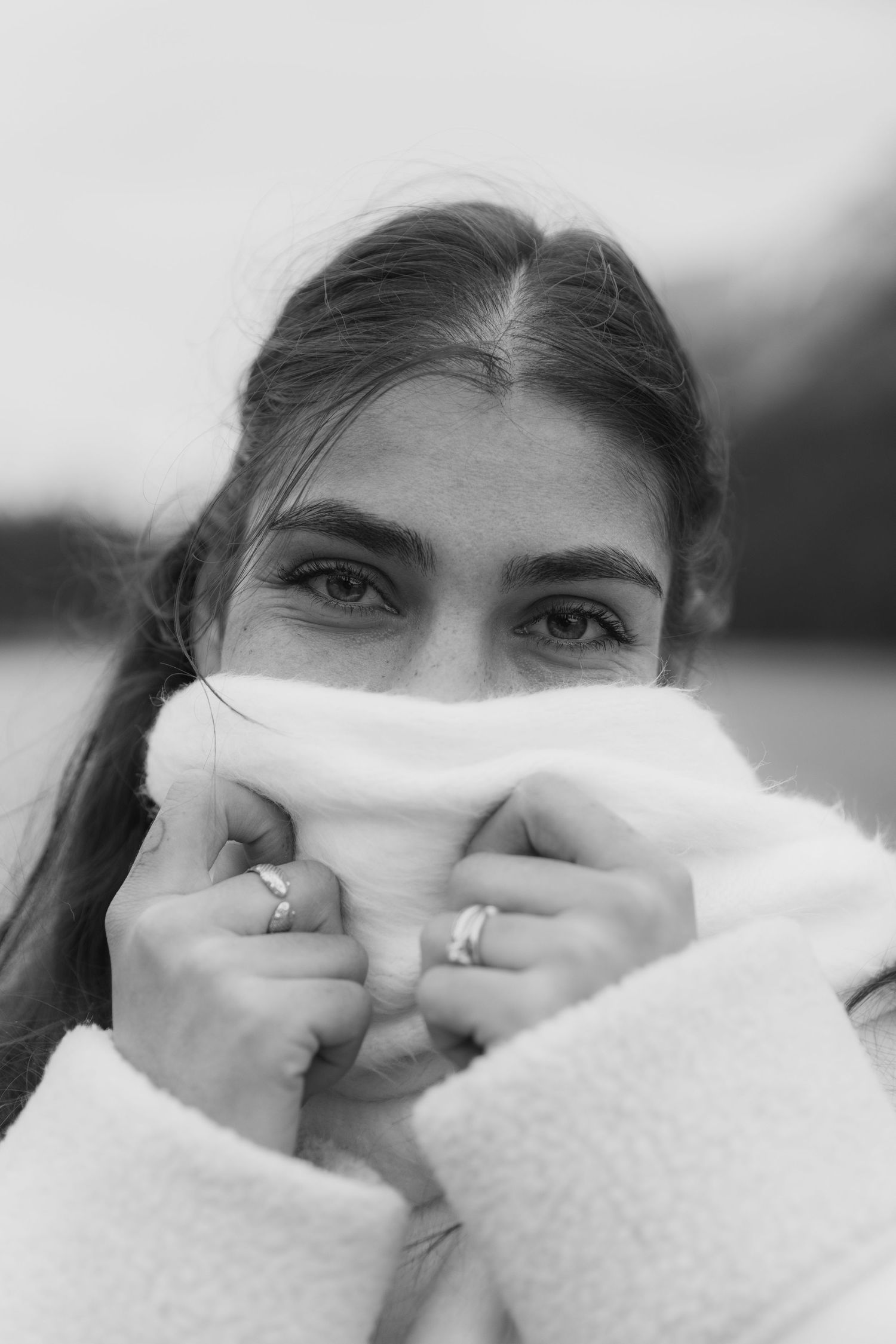 Black and white portrait of someone covering their mouth with a white sweater while showing rings on their fingers.