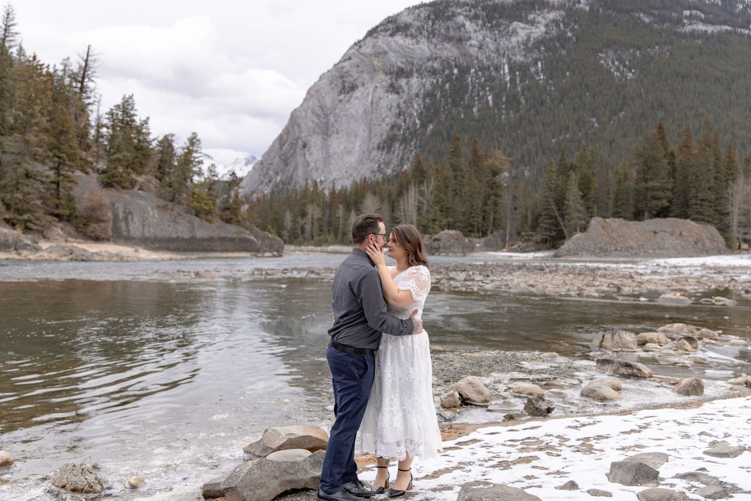A couple shares a romantic moment by the water's edge with mountains and evergreen forest in the background.