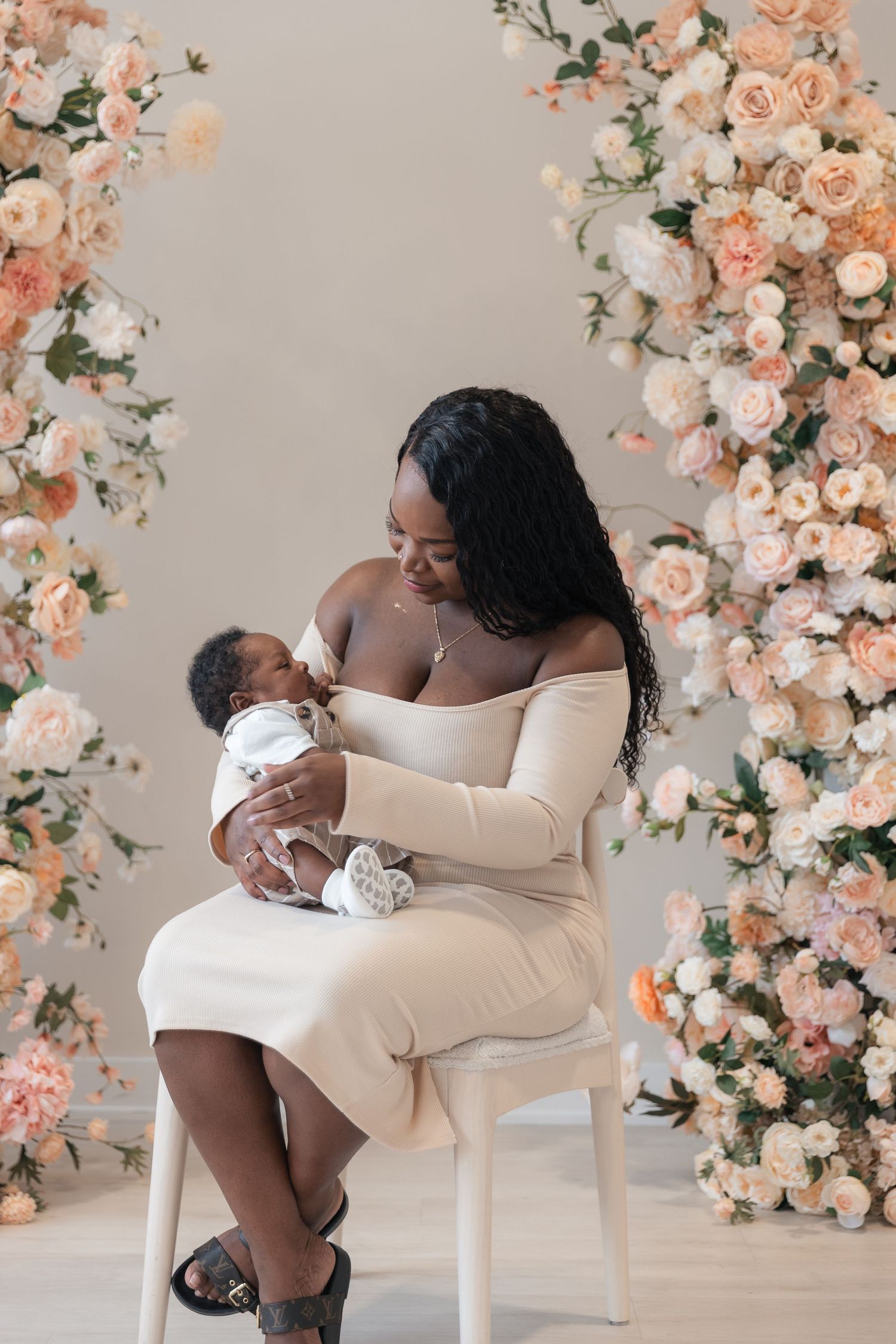 A mother in a beige off-shoulder dress cradles her newborn baby, seated before a stunning floral arch of peach roses.
