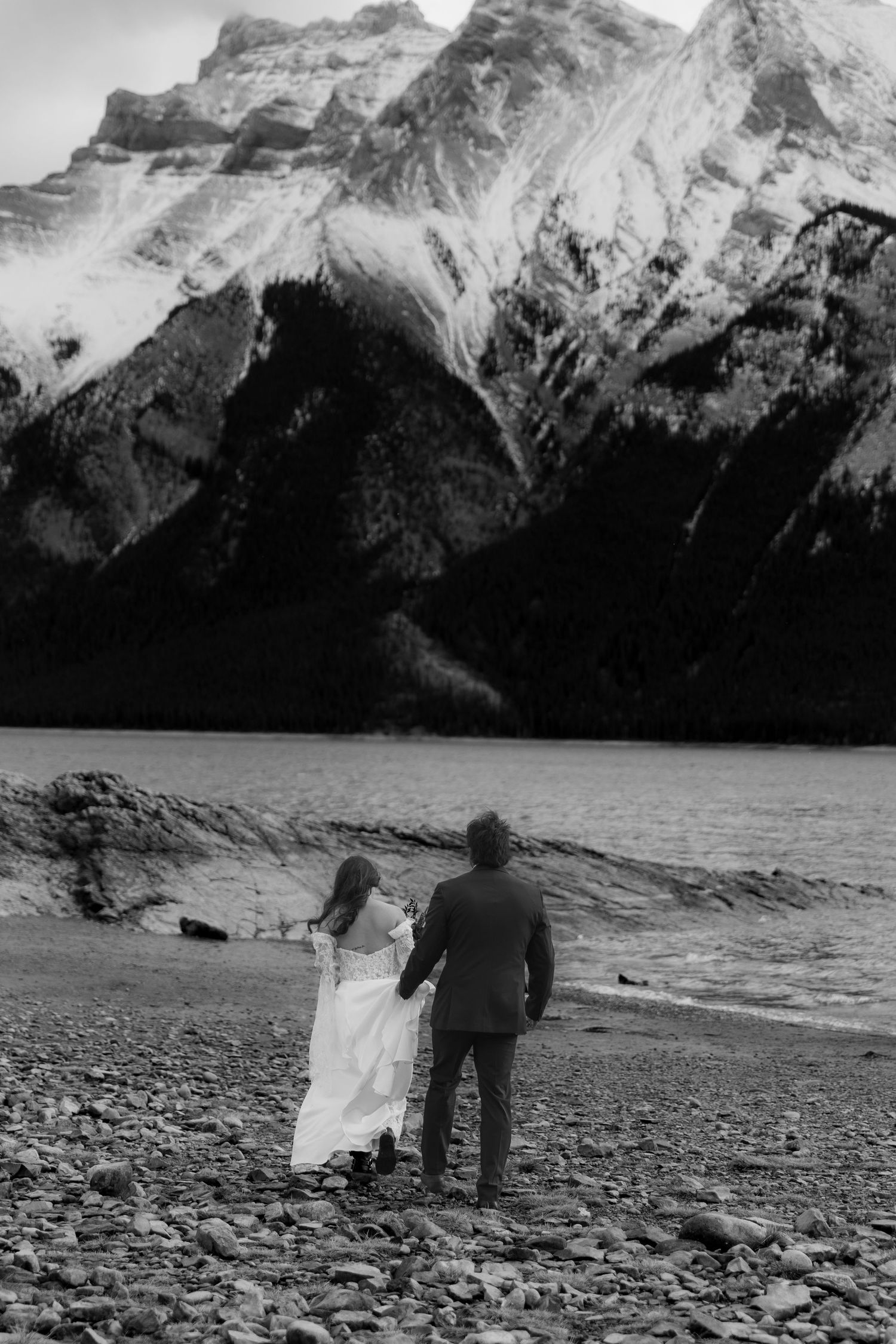 A black and white panoramic series showing people walking along a rocky mountain shoreline with towering peaks in the background.