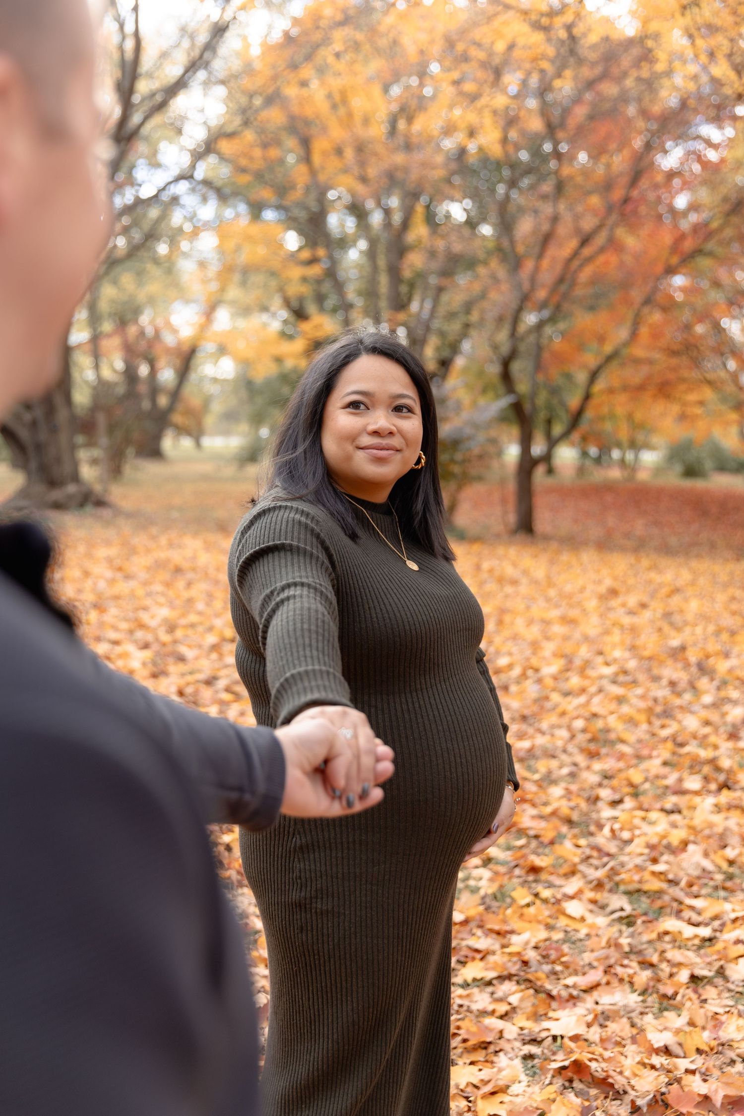 Person in dark sweater poses in autumn park with golden leaves and trees in background.