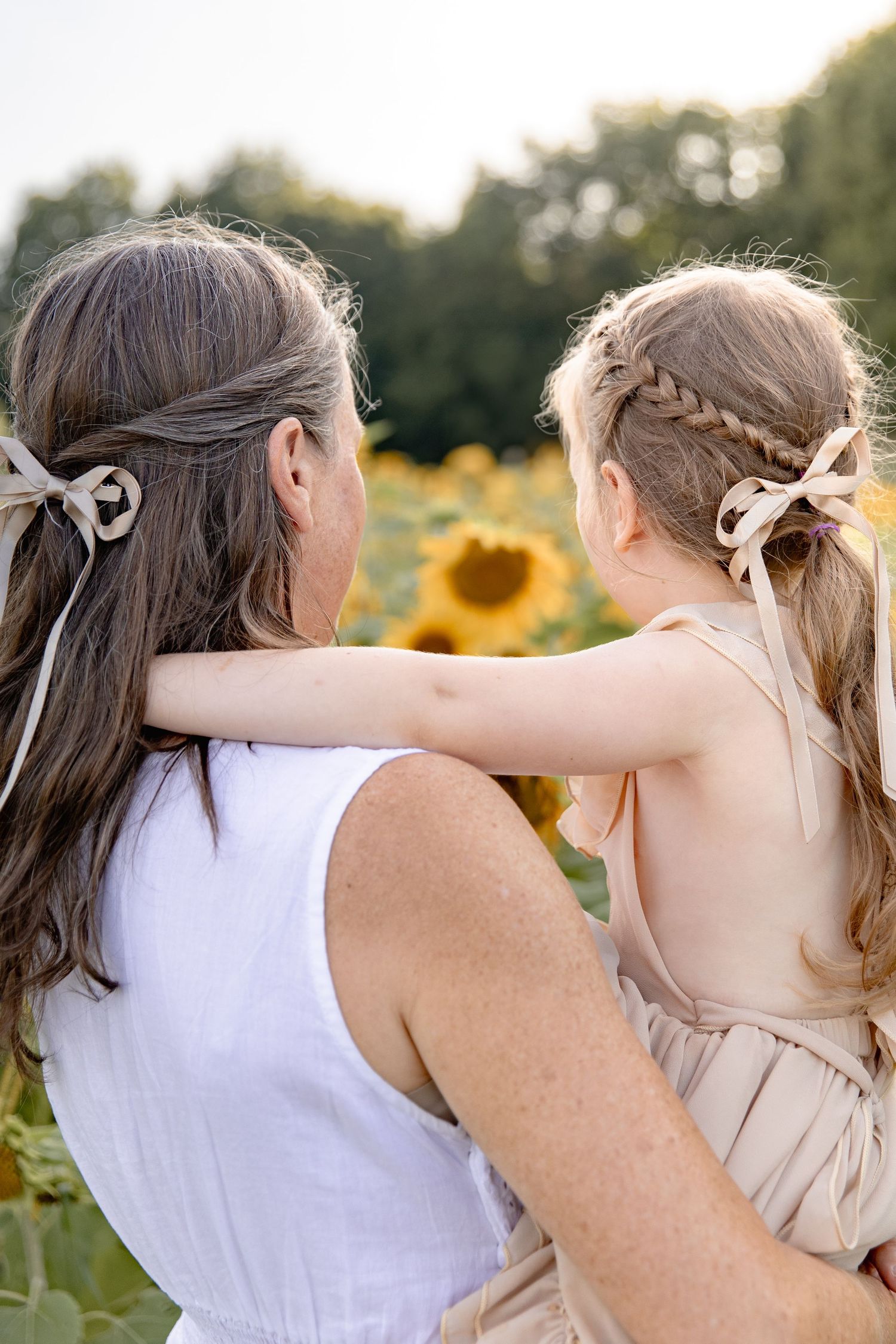 Two people in white clothing embrace while standing in a sunflower field during golden hour.