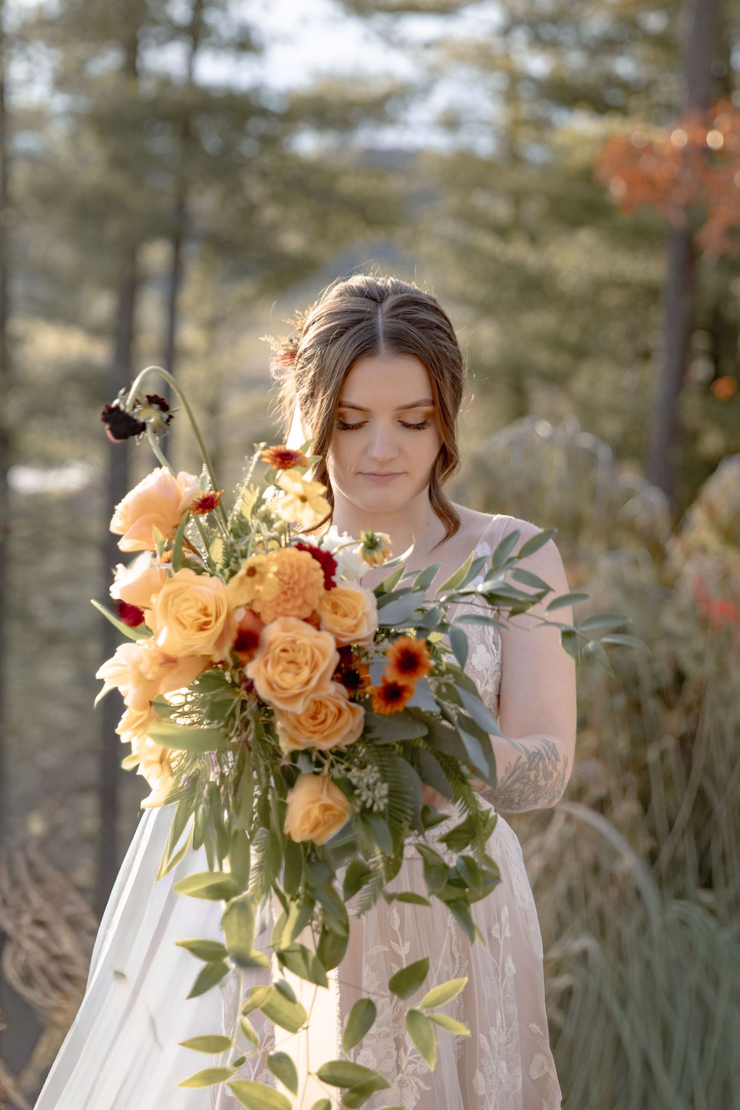 A bride in a white dress holds a romantic peach and orange bouquet with roses and greenery in golden sunlight.