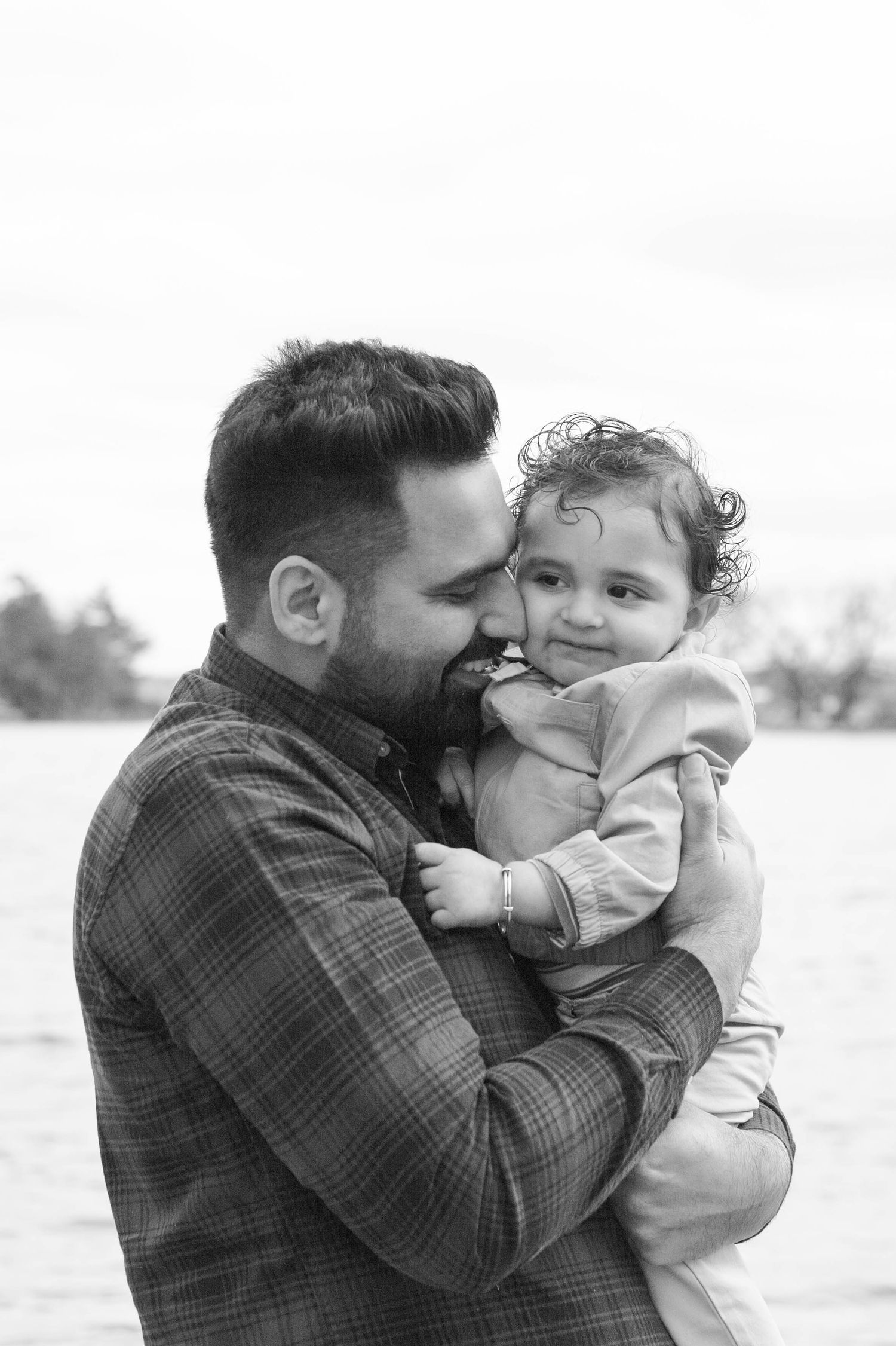 Black and white portrait of tender moment between parent and toddler against blurred outdoor background.