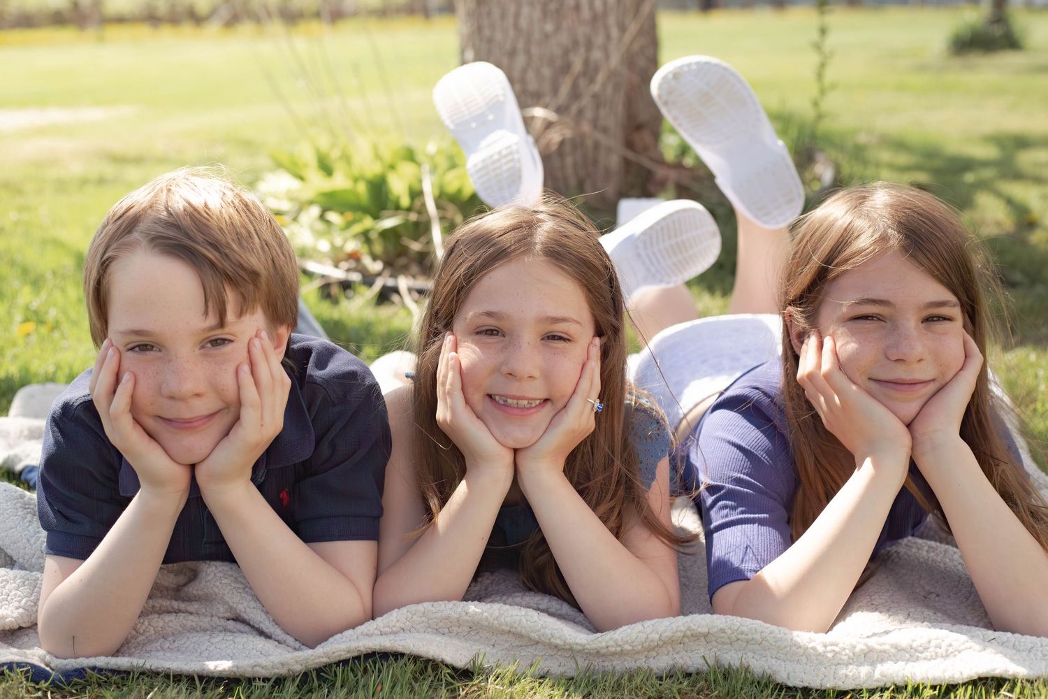 Three children lying on a blanket outdoors wearing bunny ears and resting their heads in their hands on a sunny day.