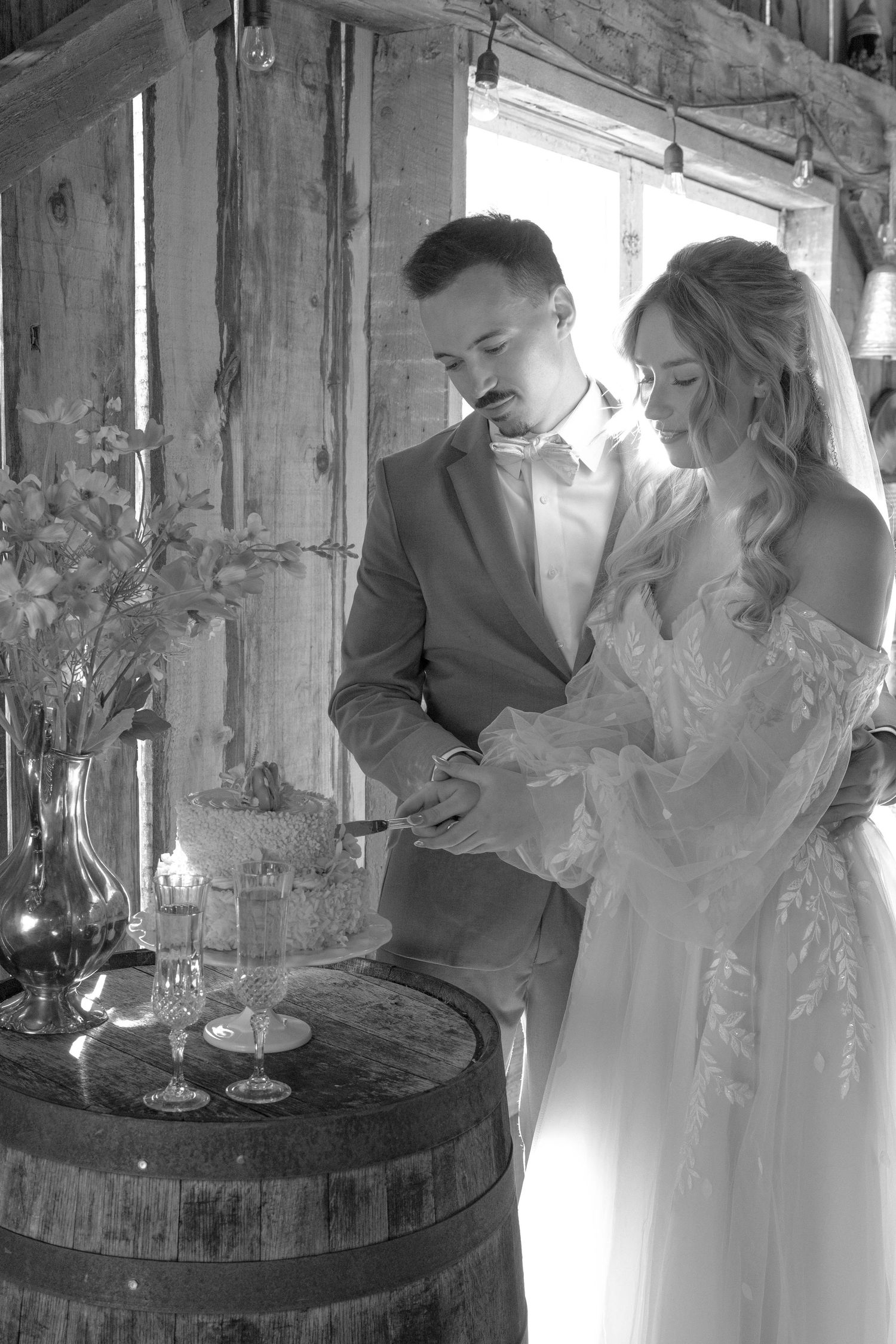 Black and white wedding photo showing couple by rustic wine barrel with wine glasses and floral arrangement in barn setting.