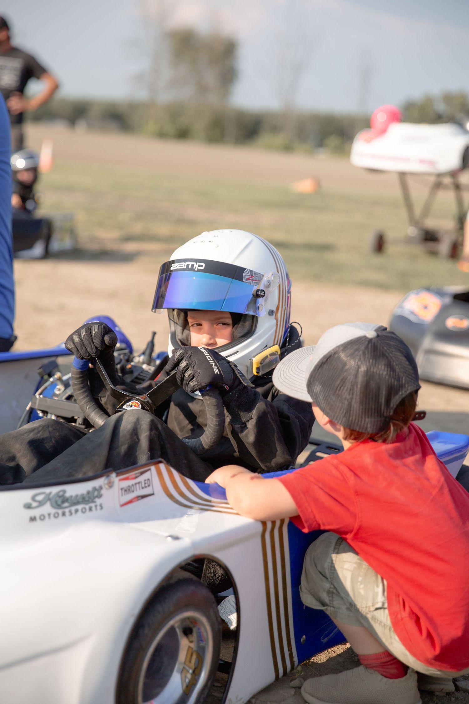 Go-kart racer in white helmet being assisted by person in red shirt at outdoor racing track.