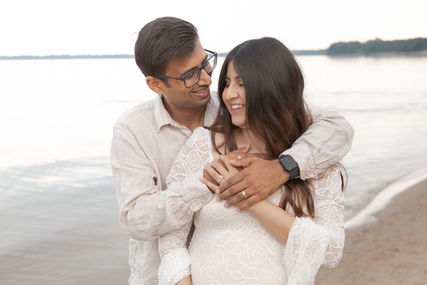 A couple embraces lovingly on a peaceful beach at sunset with calm waters in the background.