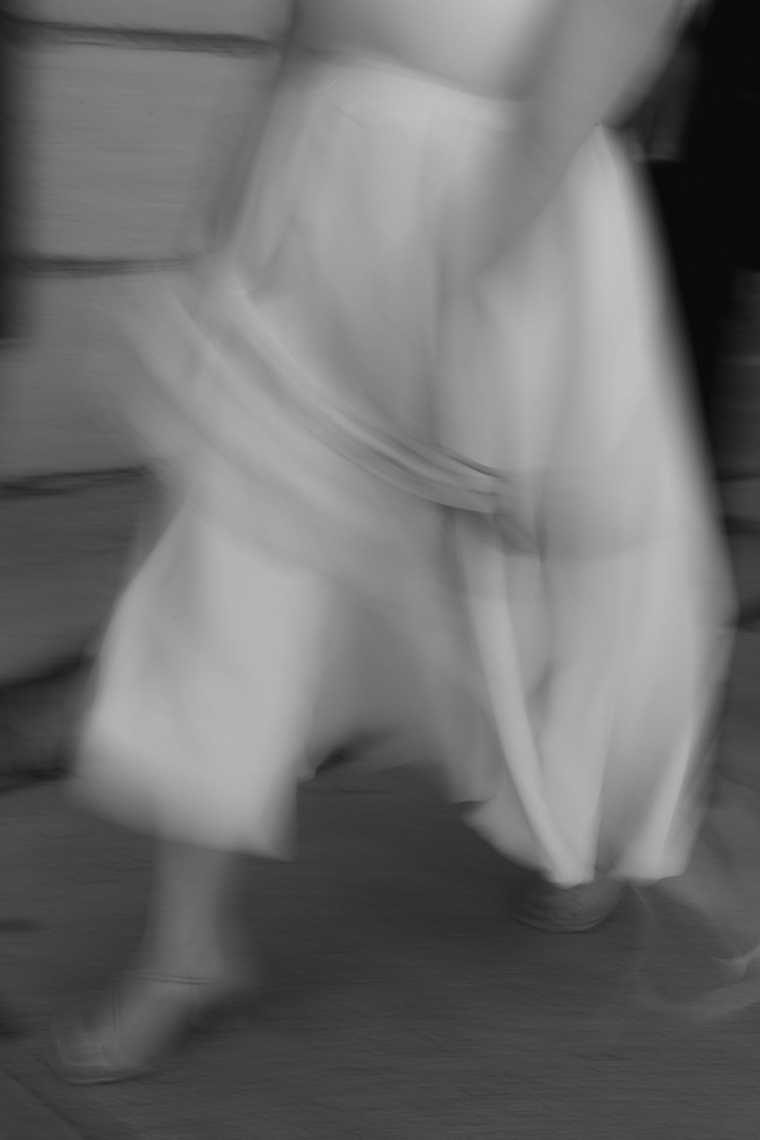 Motion-blurred black and white photograph showing the flowing movement of white fabric against a dark floor.