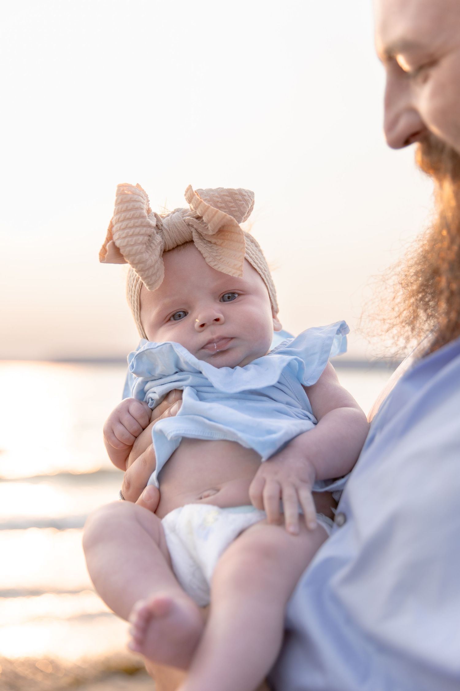 A baby wearing a light blue bow rests peacefully against someone with a beard in a tender outdoor sunset moment.