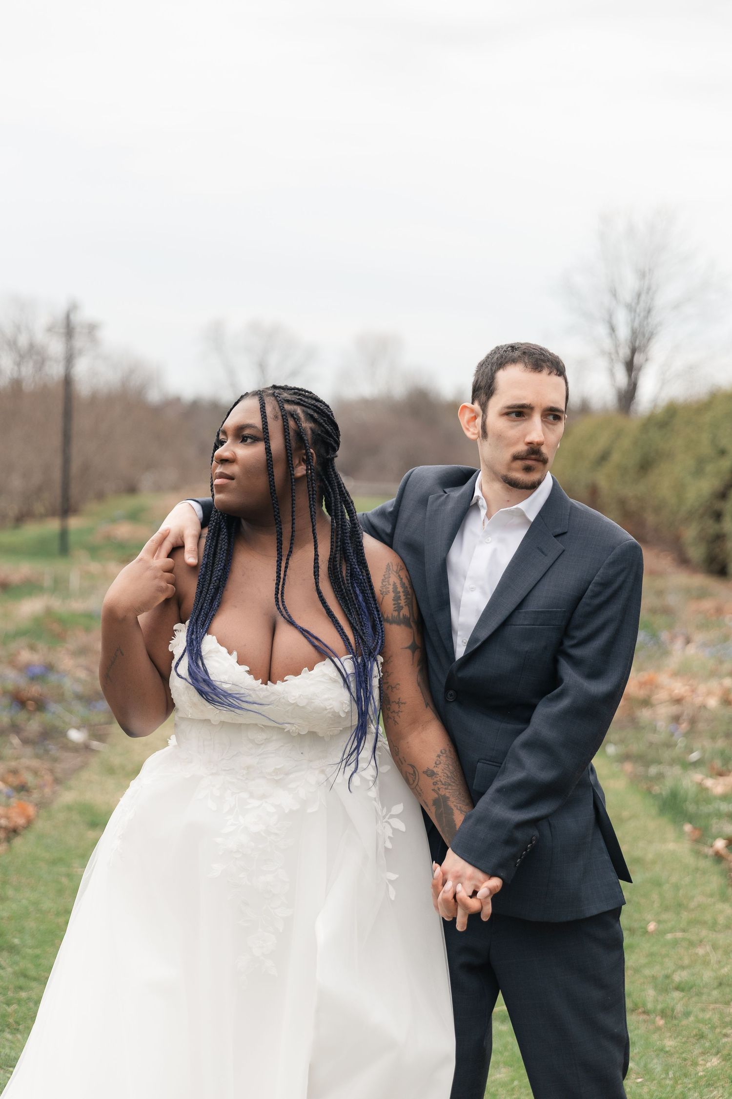 Interracial couple in wedding attire holding hands outdoors, bride in white lace gown with blue braids, groom in dark suit.