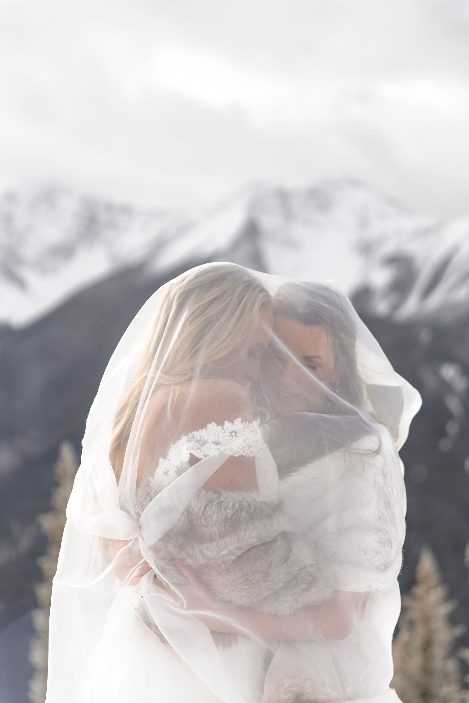 A bride wrapped in a delicate white veil stands against a backdrop of snow-capped mountains on her wedding day.