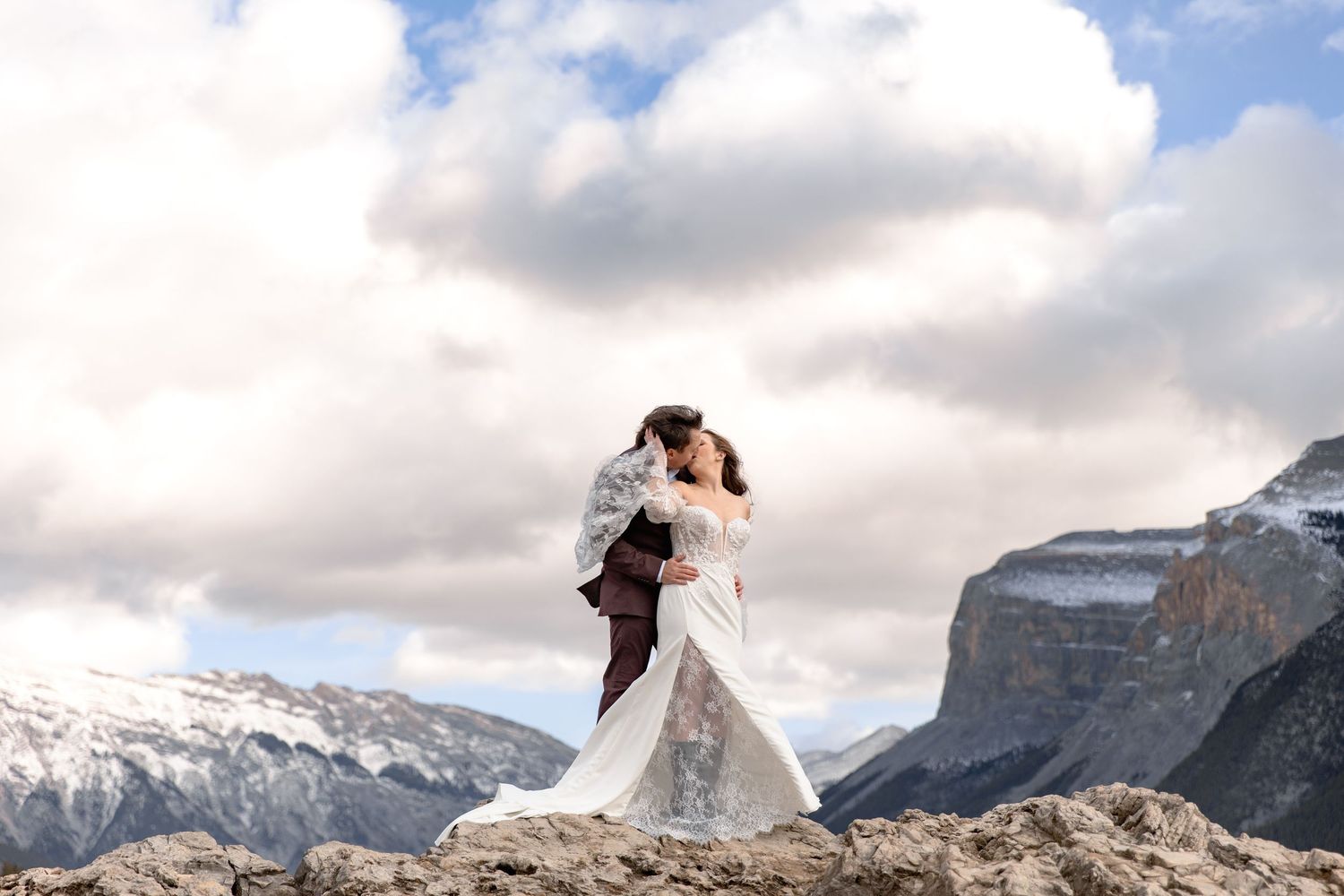 A couple poses in wedding attire against a dramatic mountain backdrop with cloudy skies and snow-capped peaks.