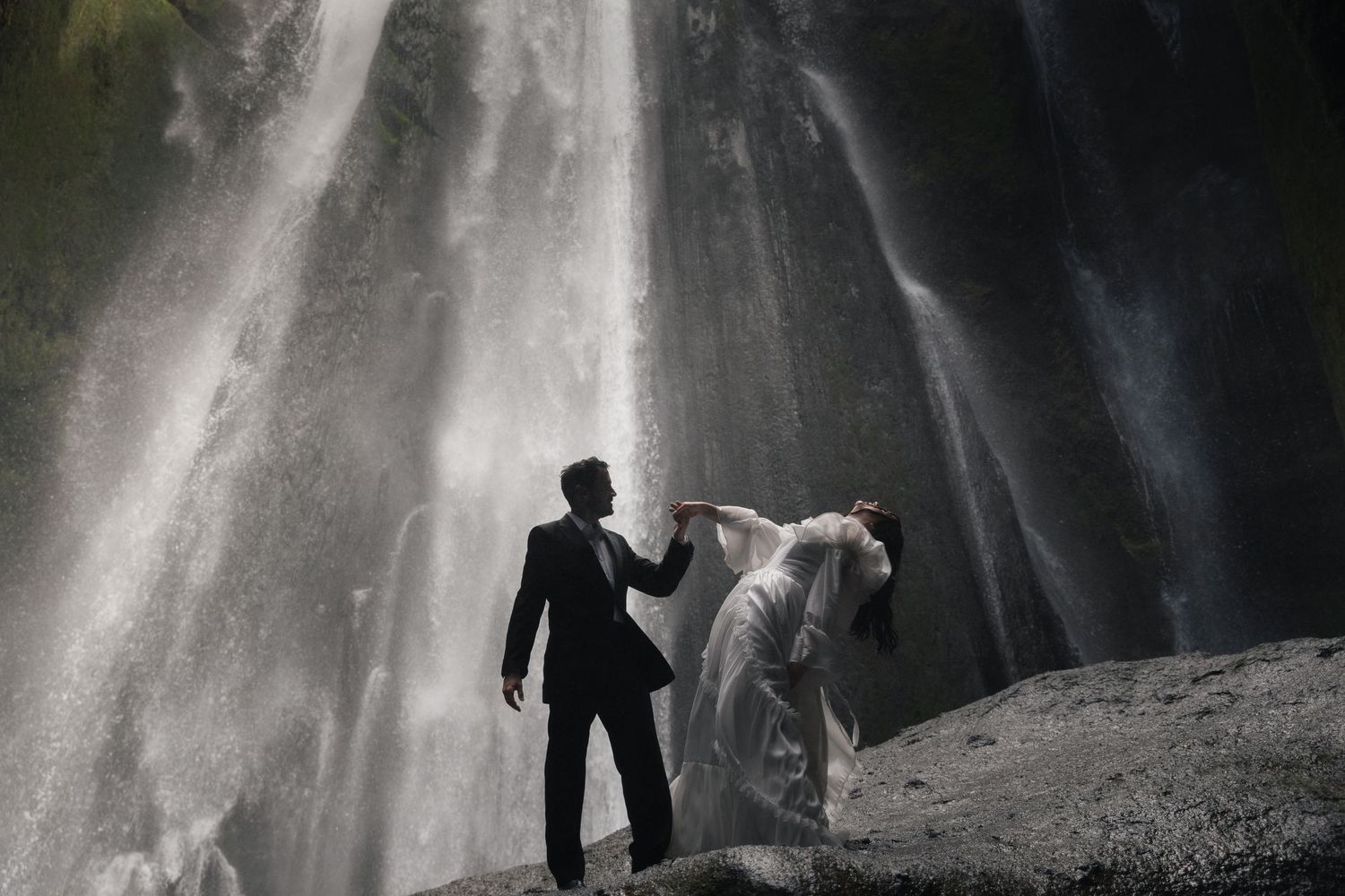 Bride and groom pose romantically before a stunning waterfall, with mist and dramatic light surrounding them.