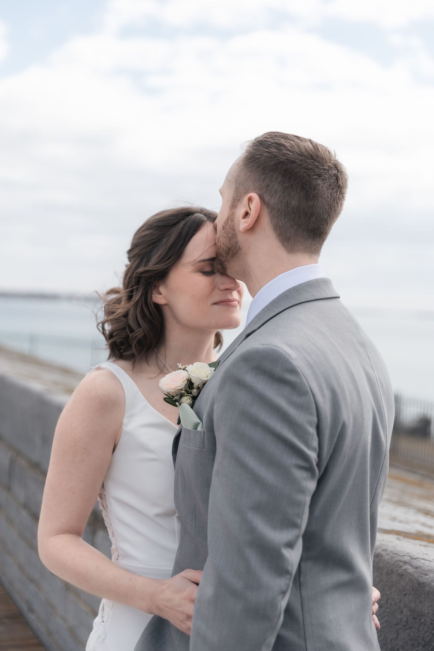 Bride and groom share a tender kiss outdoors near a waterfront, with the groom in a gray suit and bride in a white dress.