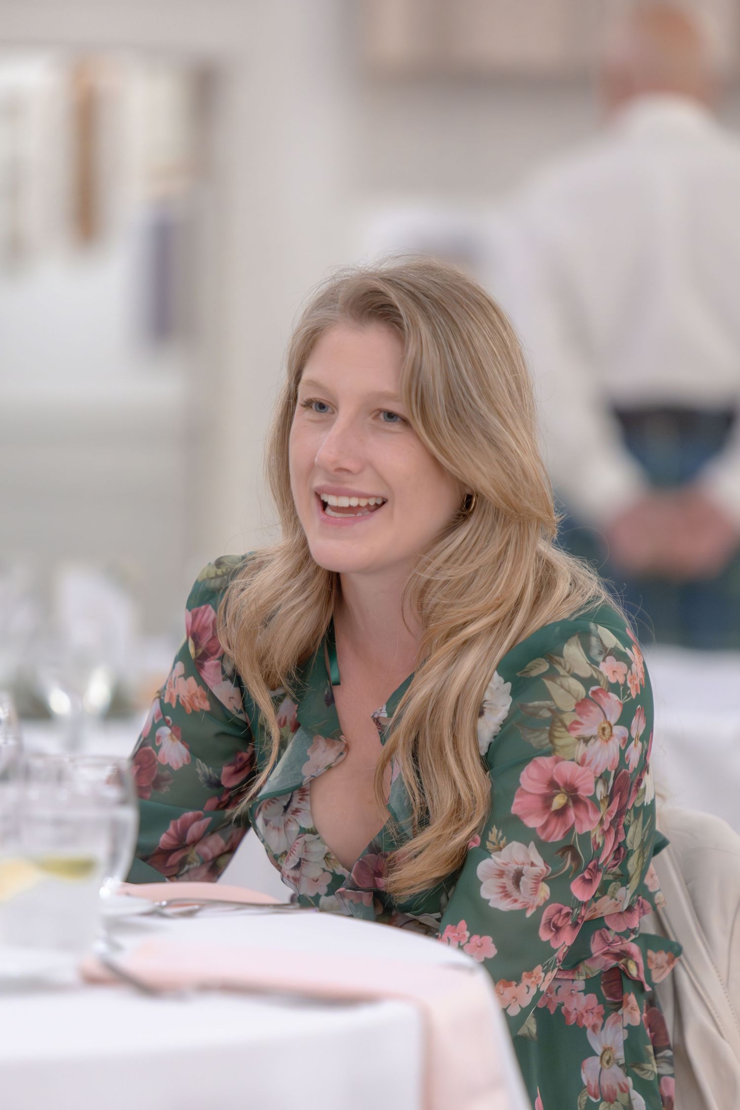 A person in a green floral dress smiles while seated at a dining table.