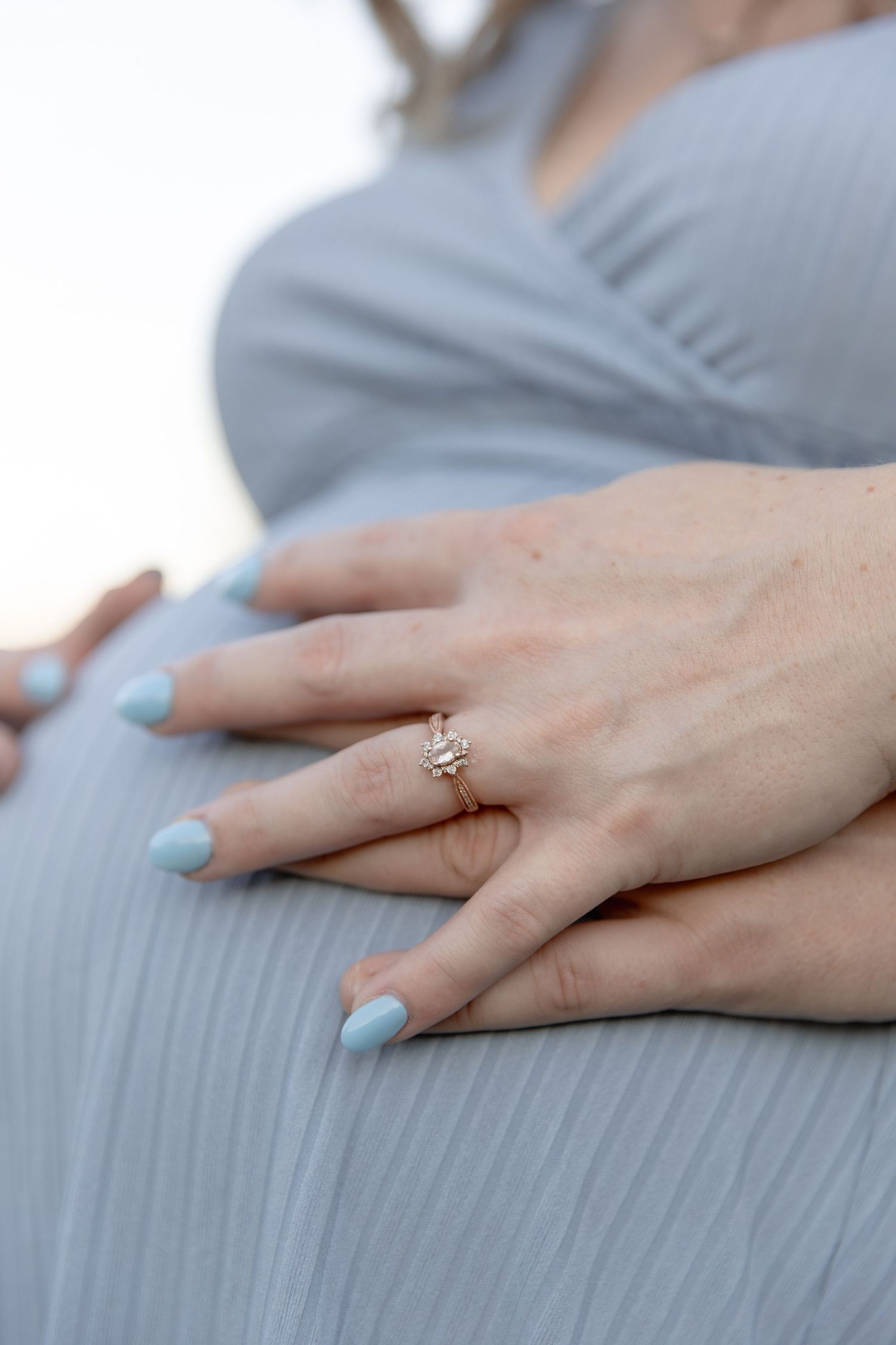 Close-up of a hand with light blue nail polish and a delicate ring resting on a gray fabric surface.