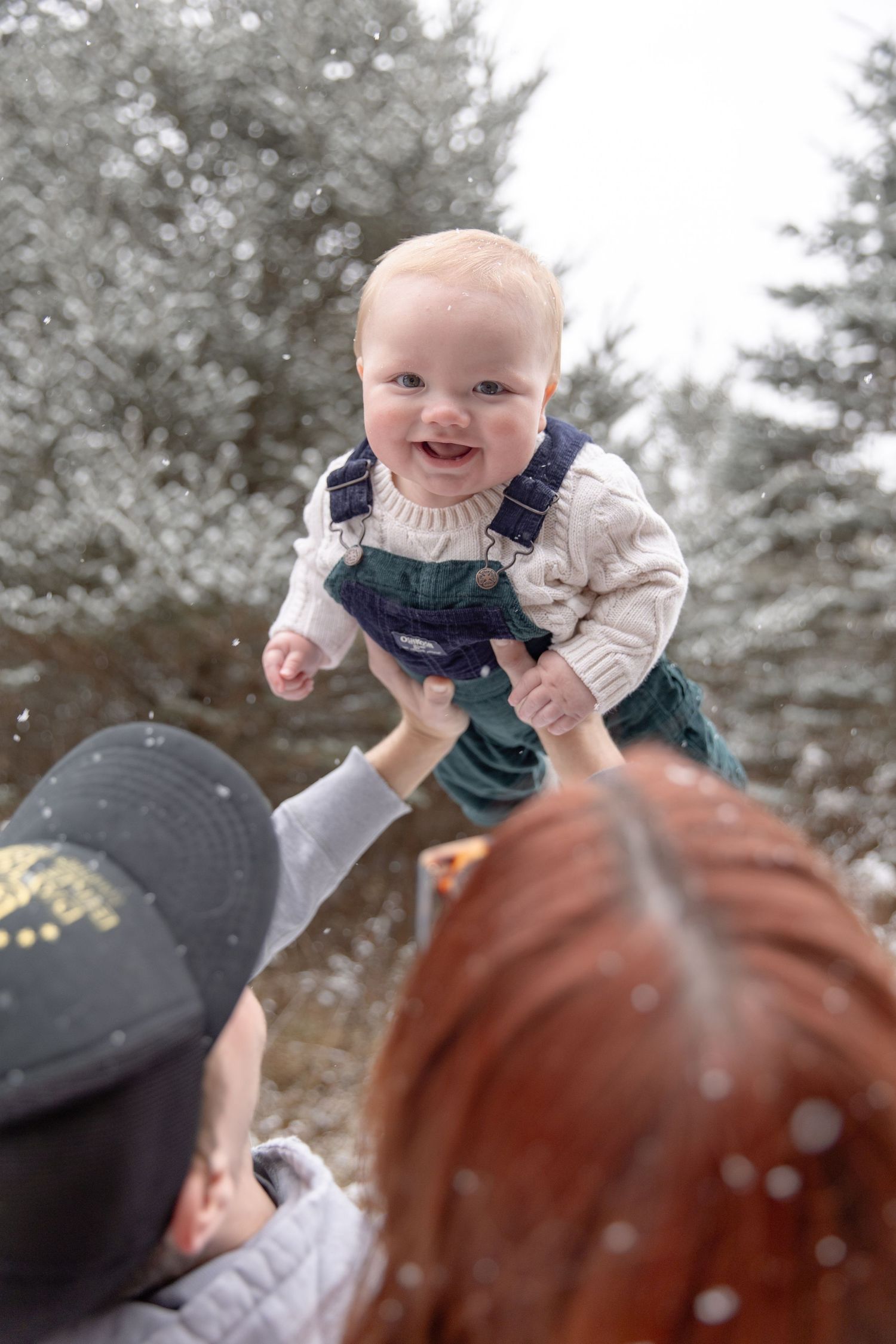 A series of joyful moments captured in winter as a baby is lifted up high against a snowy evergreen forest backdrop.