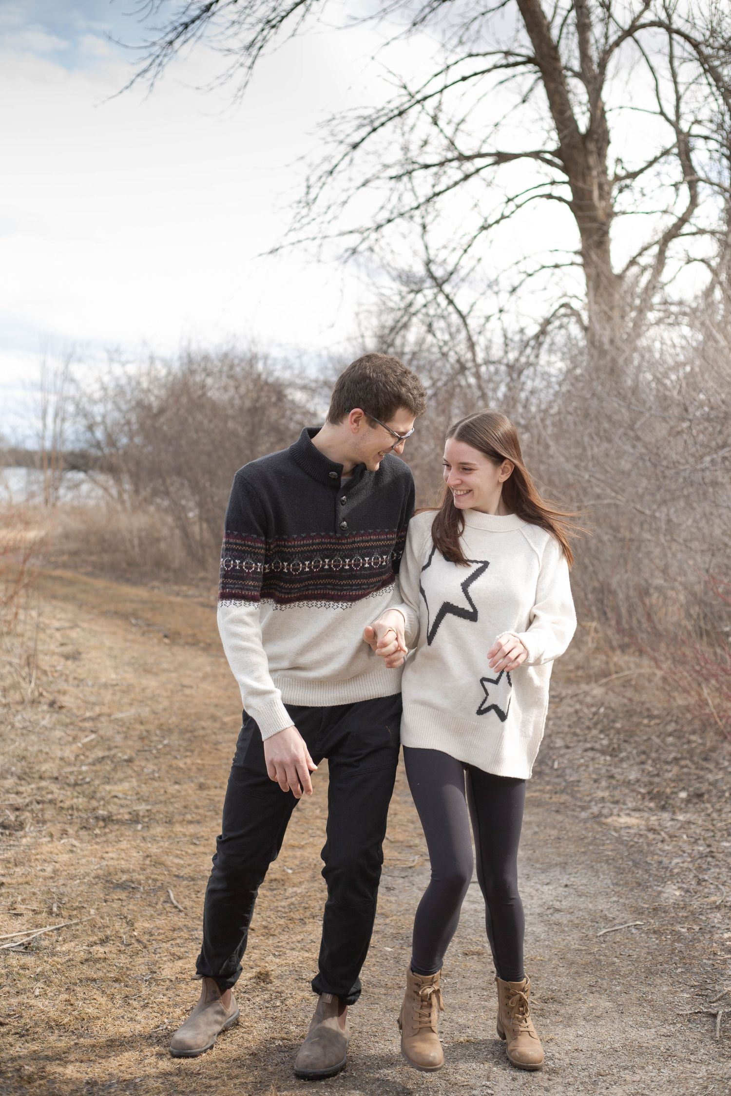 A couple in cozy winter sweaters walking together on a dirt path through bare trees on an autumn day.