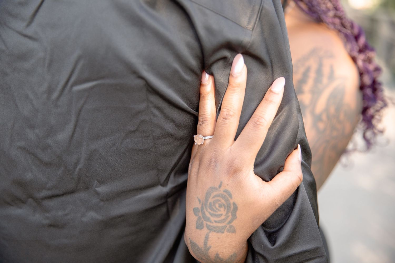 Hand with rose tattoo rests on black fabric, wearing a silver ring on finger.