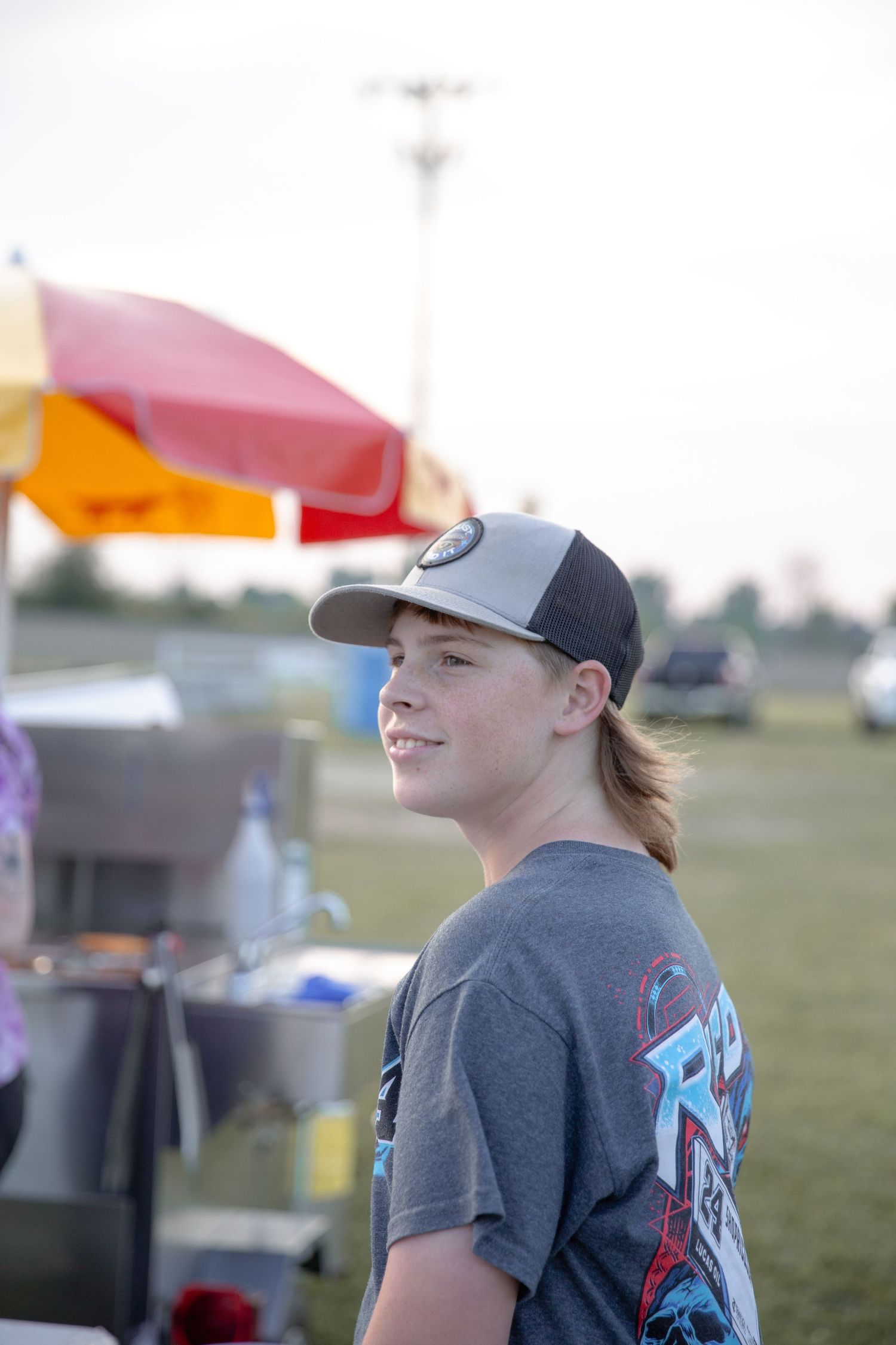 A player in a grey t-shirt and baseball cap stands near tents at a sports field during an outdoor event.