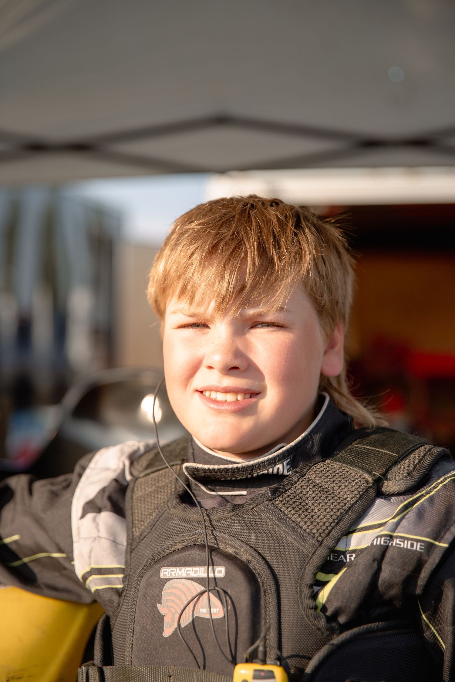 Rider in a black protective motorsport suit looks ahead while illuminated by warm sunlight.