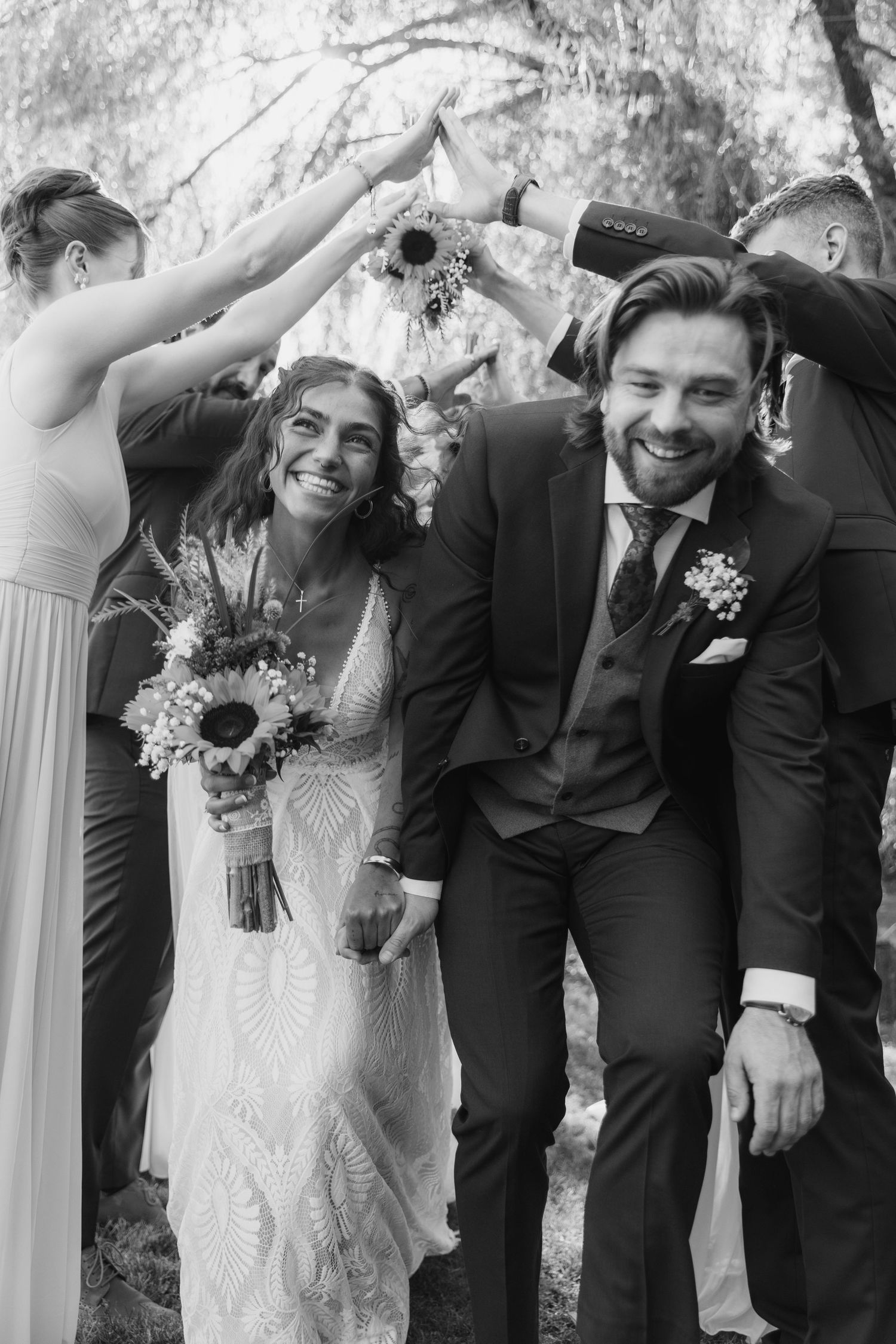 Wedding party in black suits and white dresses pose playfully in a line against a light background.