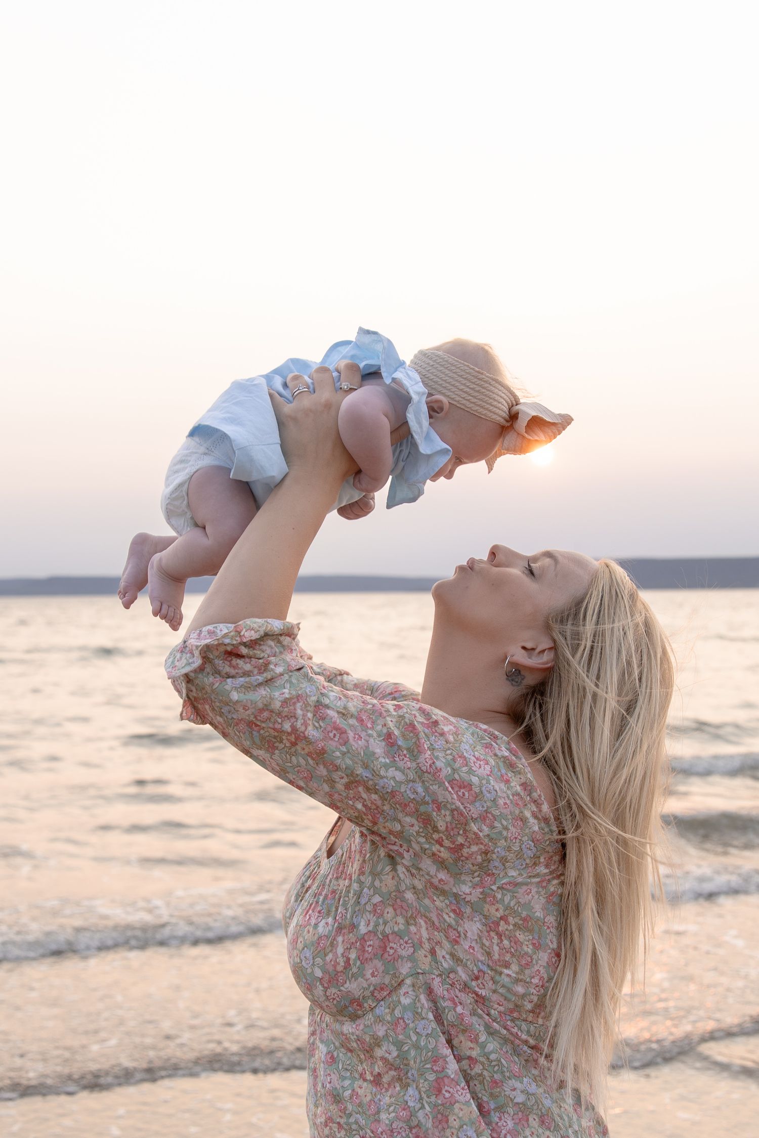 Woman in floral dress lifts baby high into air at sunset on beach with ocean in background.