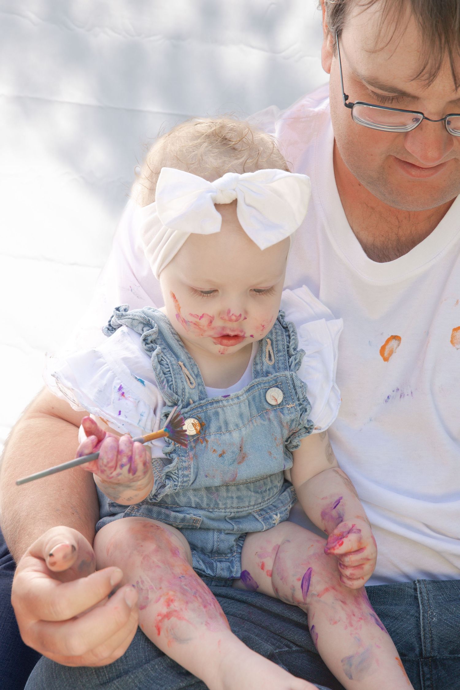 A baby in denim overalls gets messy with colorful paint while being held by someone in a white shirt.