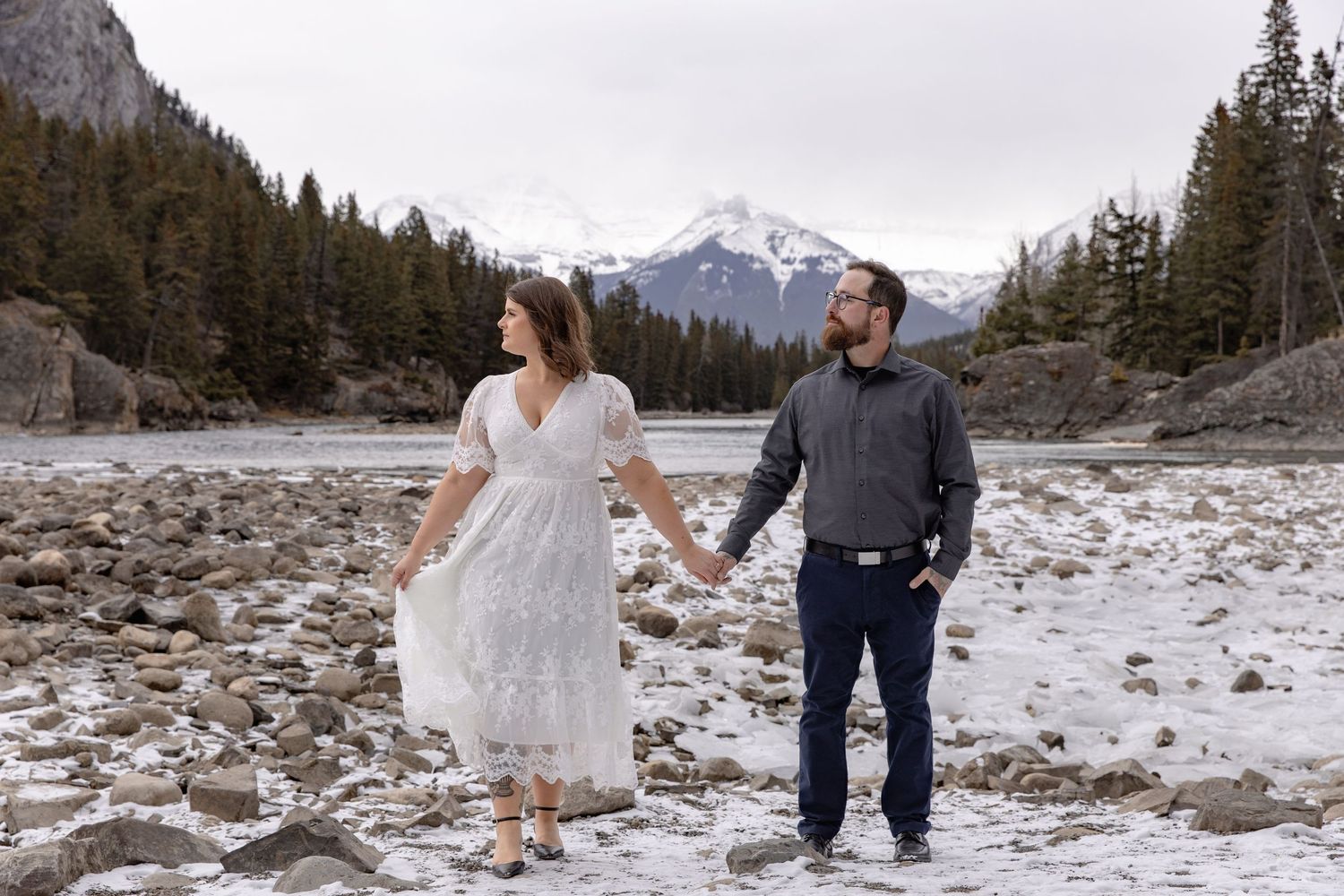 A couple in wedding attire walks hand in hand through a scenic mountain landscape with snow-capped peaks in the background.