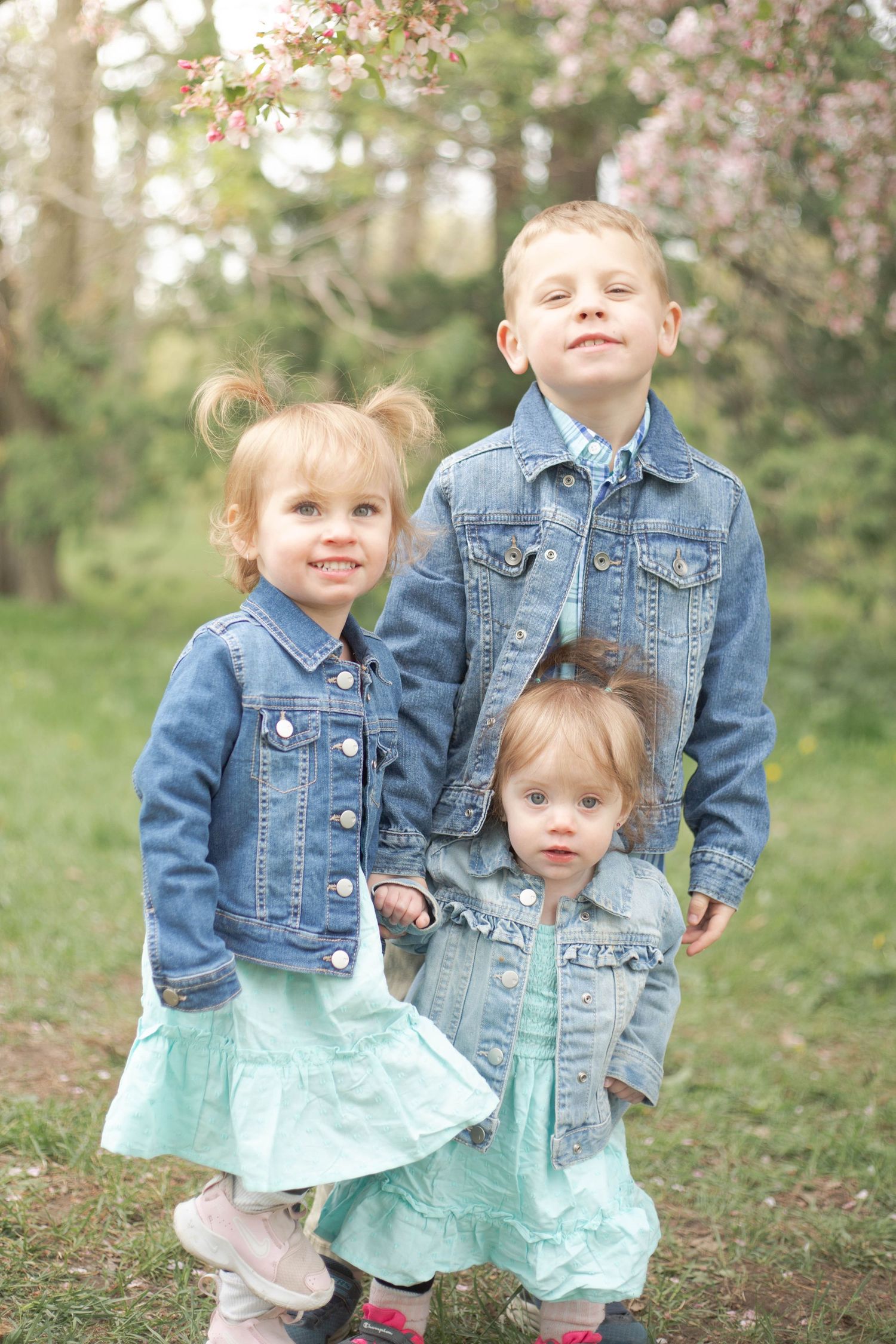 Two children in denim jackets and mint green dresses pose playfully in a spring garden setting.