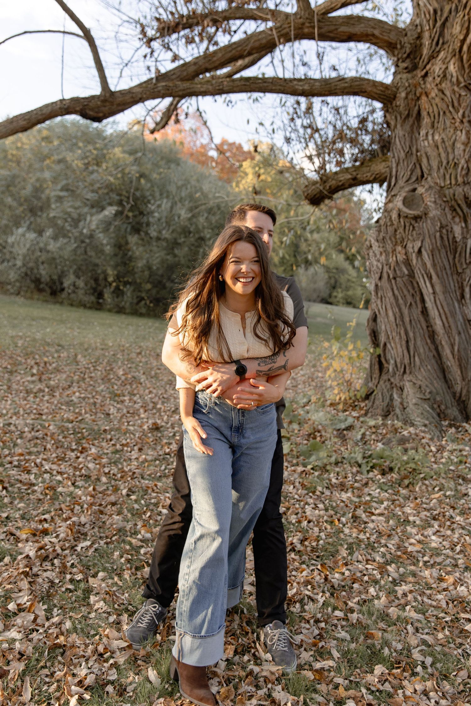 Couple embracing and laughing together in autumn forest setting with fallen leaves on ground during outdoor photoshoot.
