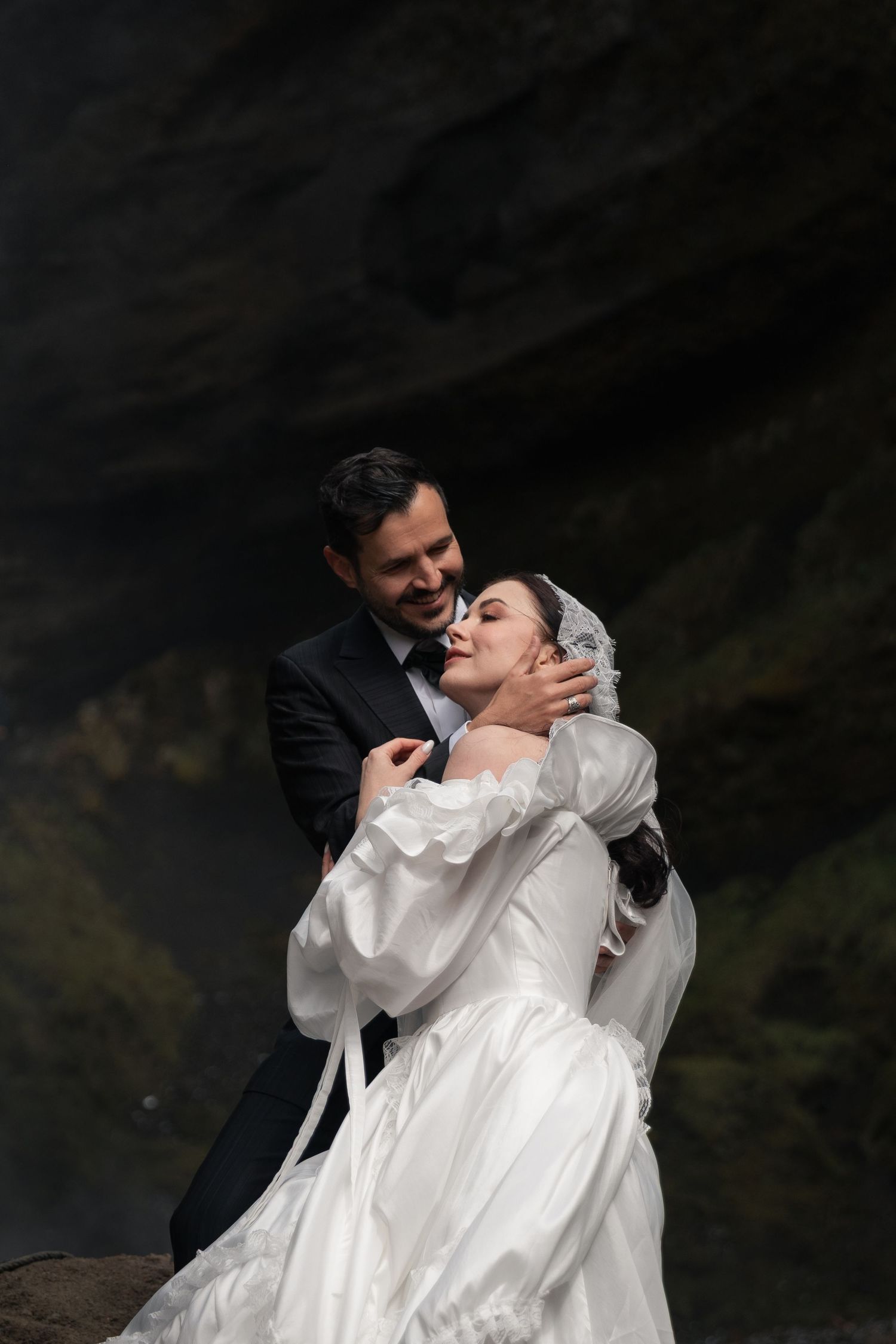 A groom in a dark suit embraces his smiling bride in a white gown and veil against a rocky backdrop.
