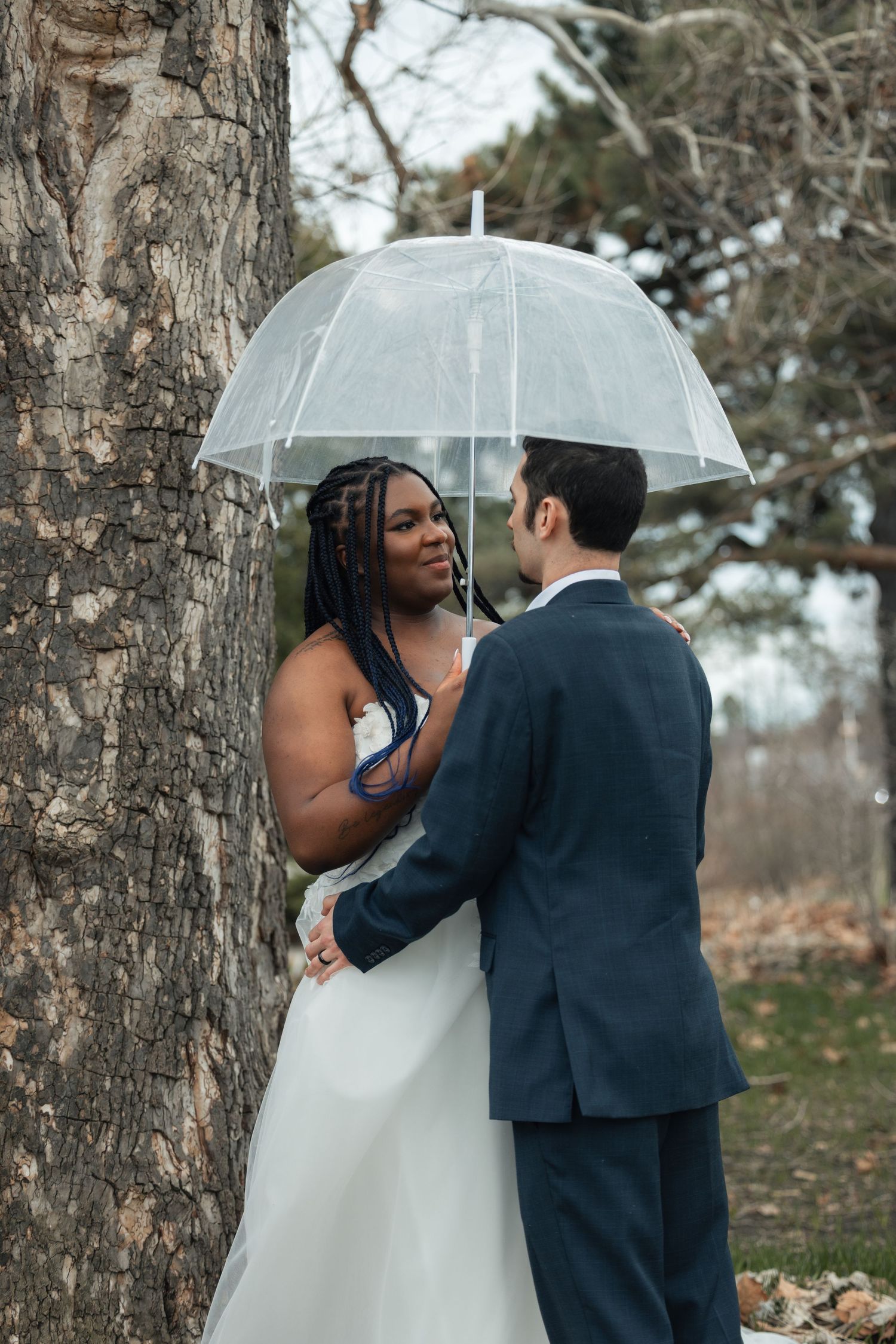 Bride and groom share a romantic moment under a clear umbrella by a tree on their rainy wedding day.
