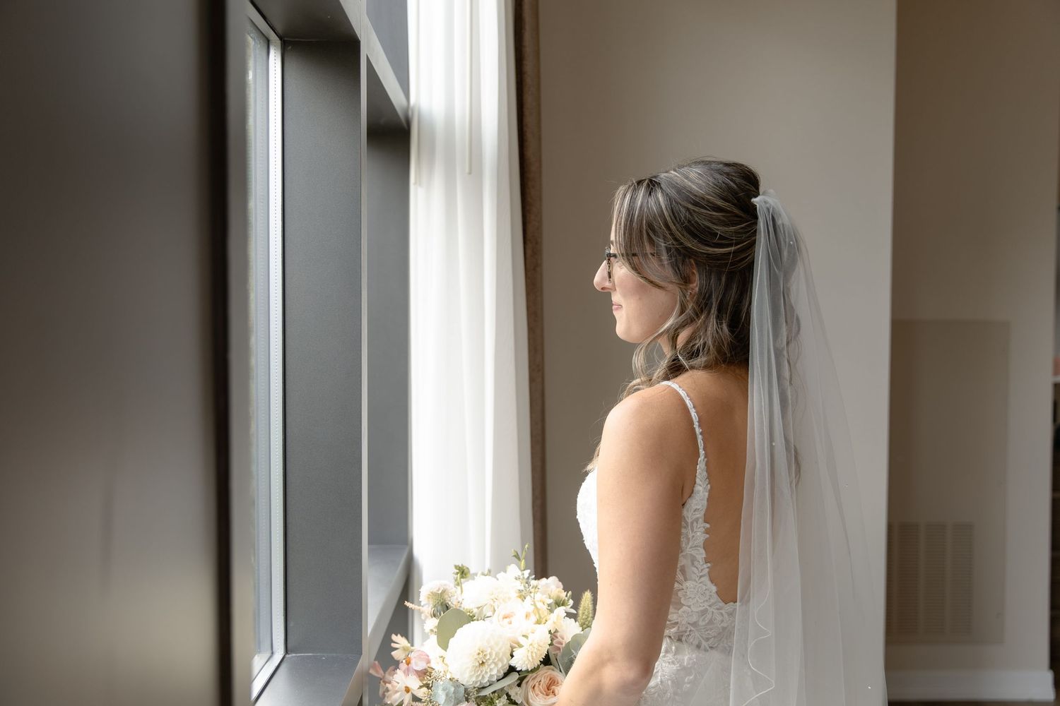 A bride in a white wedding gown holds a bouquet while looking out a large window in an elegant room.